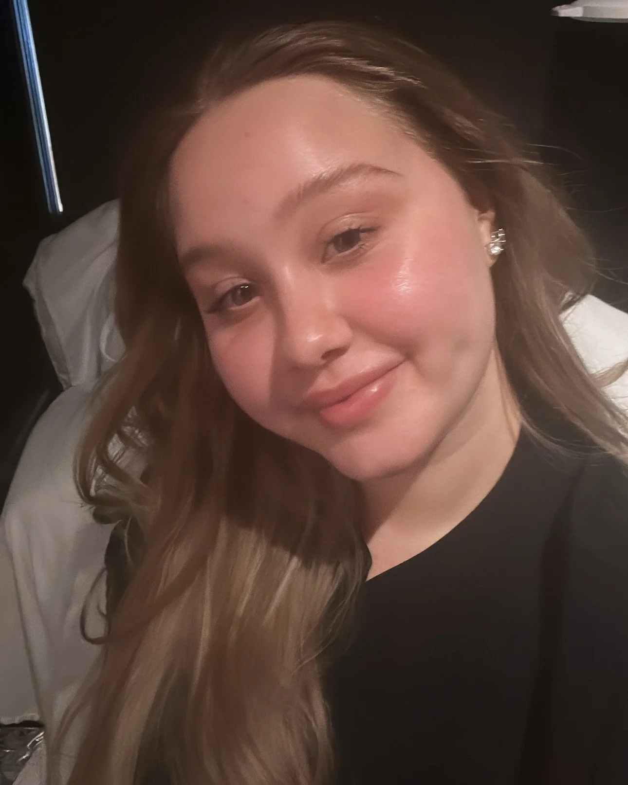 A woman with long, wavy hair, smiling, wearing pearl earrings, lying on a bed with a white pillow in the background.