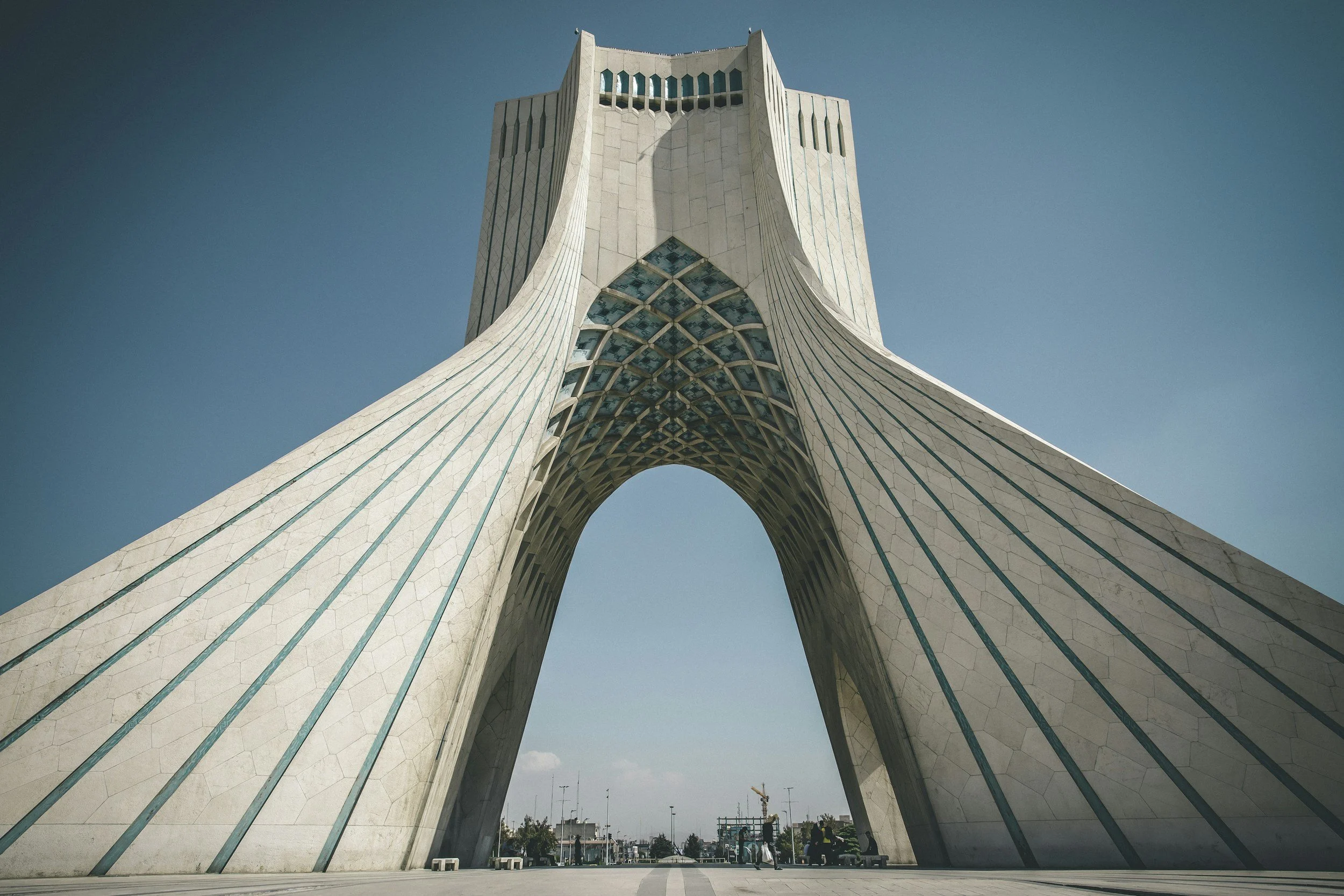Low-angle view of the Azadi Tower in Tehran, Iran, showcasing its modern, arching architectural design against a clear blue sky.