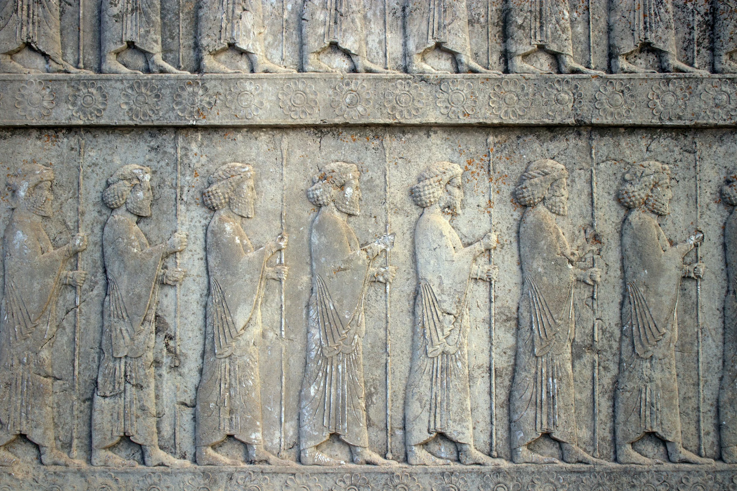 Ancient stone relief depicting a row of seven male figures in profile, each holding a staff, with elaborate hairstyles and draped clothing.