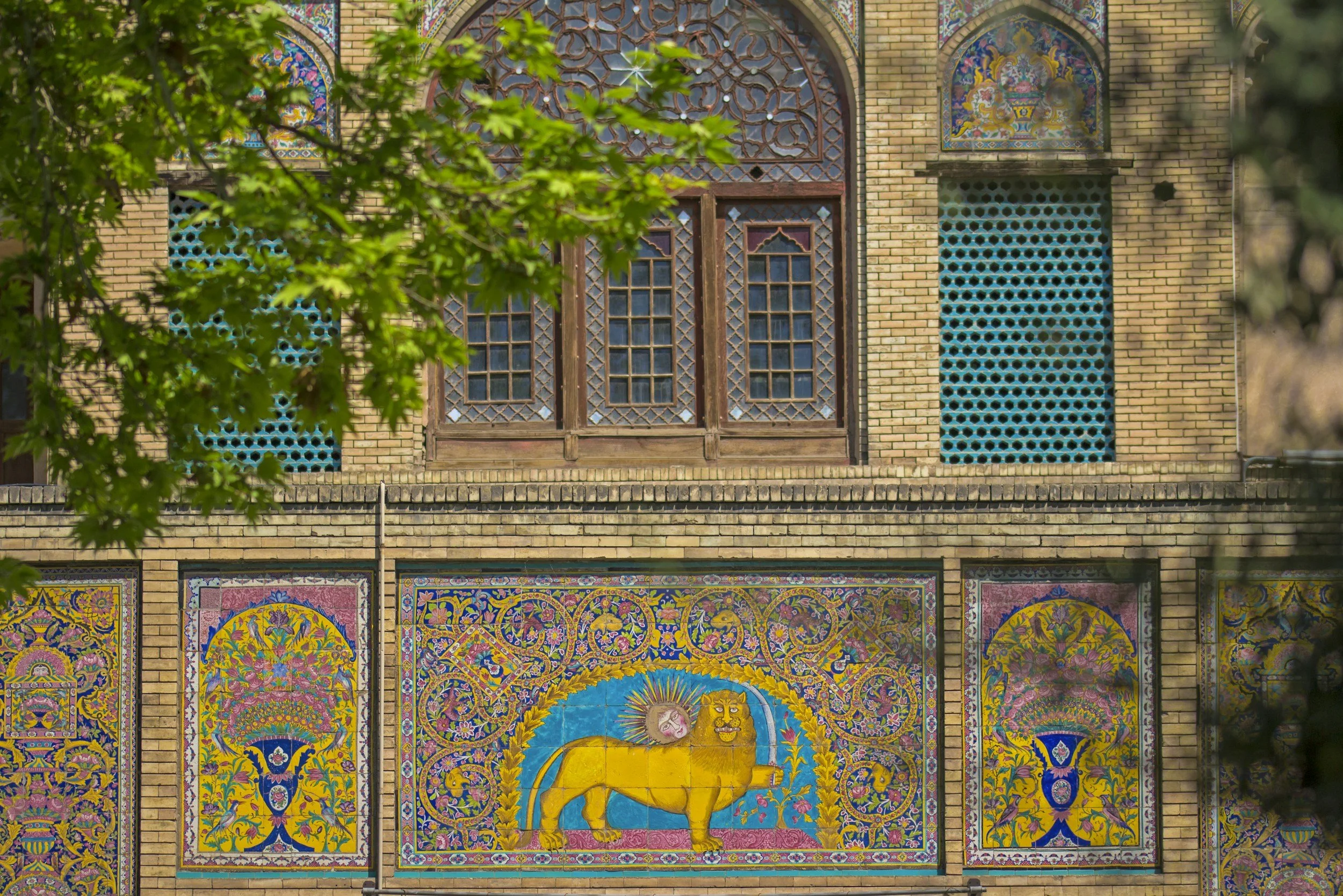Colorful mosaic on the side of a brick building depicting a yellow lion with a matthew and a sun behind it, surrounded by intricate floral patterns.