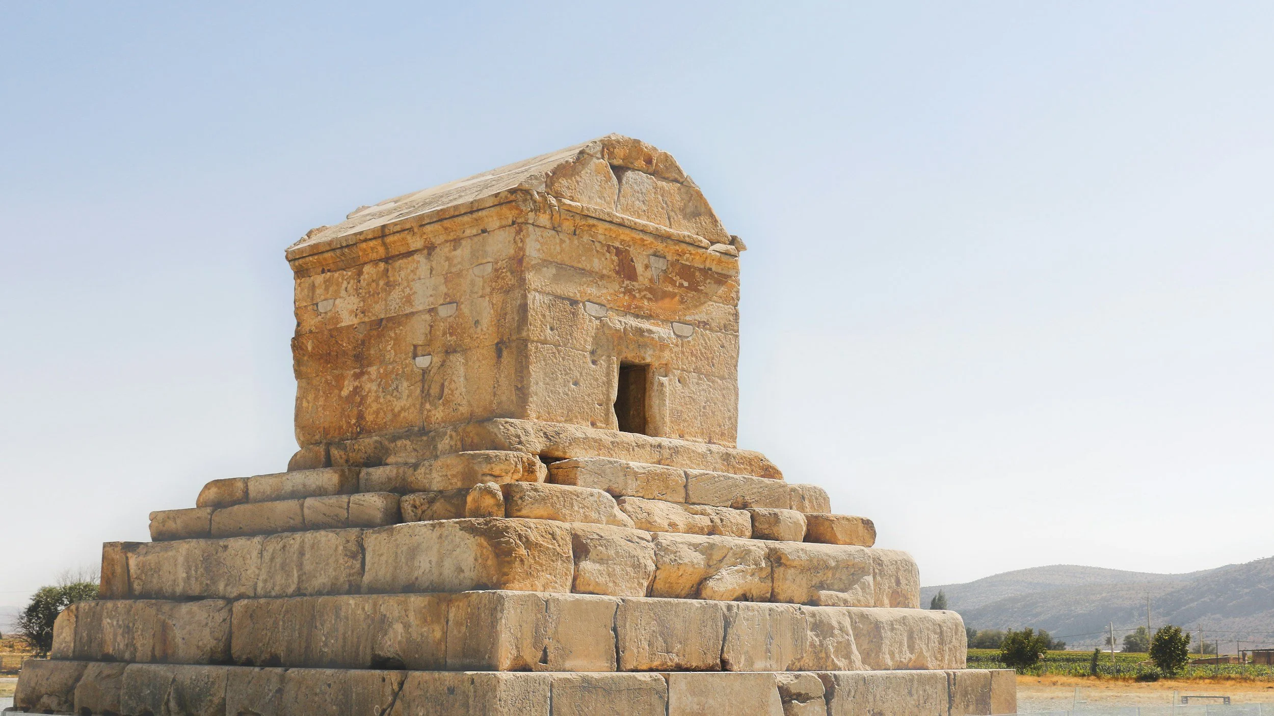 Ancient stone structure with a stepped base and small opening, set in a landscape with mountains in the background.