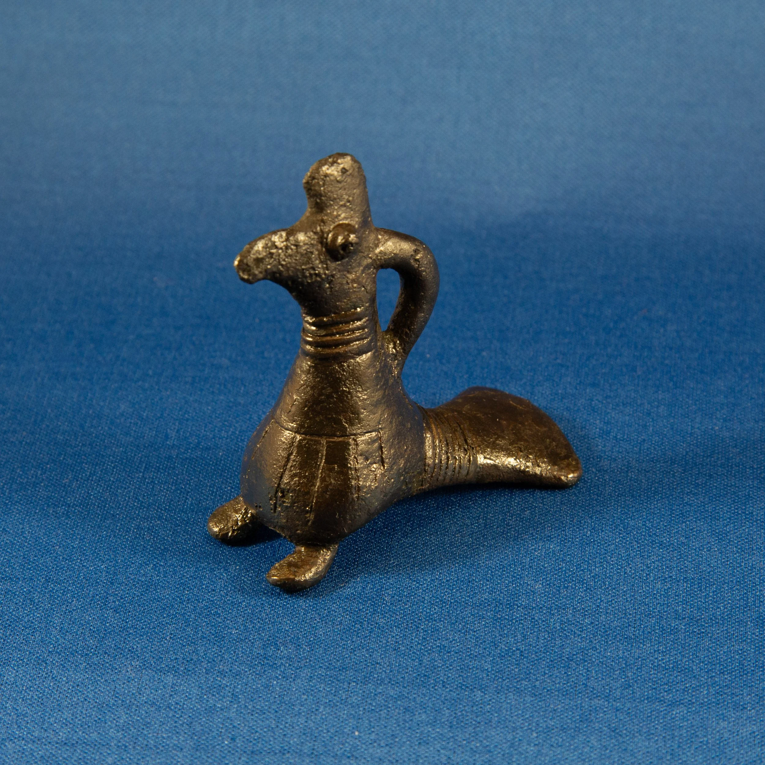 Small metallic figurine of a creature with a fish-like body, a head with a snout, and a small rounded eye, placed on a blue textured surface.