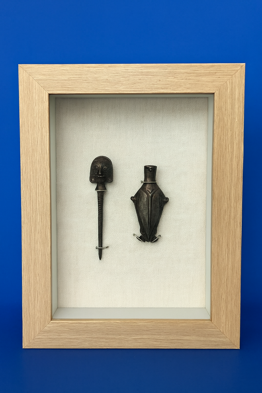Two ancient tribal artifacts mounted in a display case with a white fabric background and a wooden frame against a blue wall. One artifact appears to be a carved wooden staff with a face at the top, and the other is a carved animal figure, possibly a frog or toad.