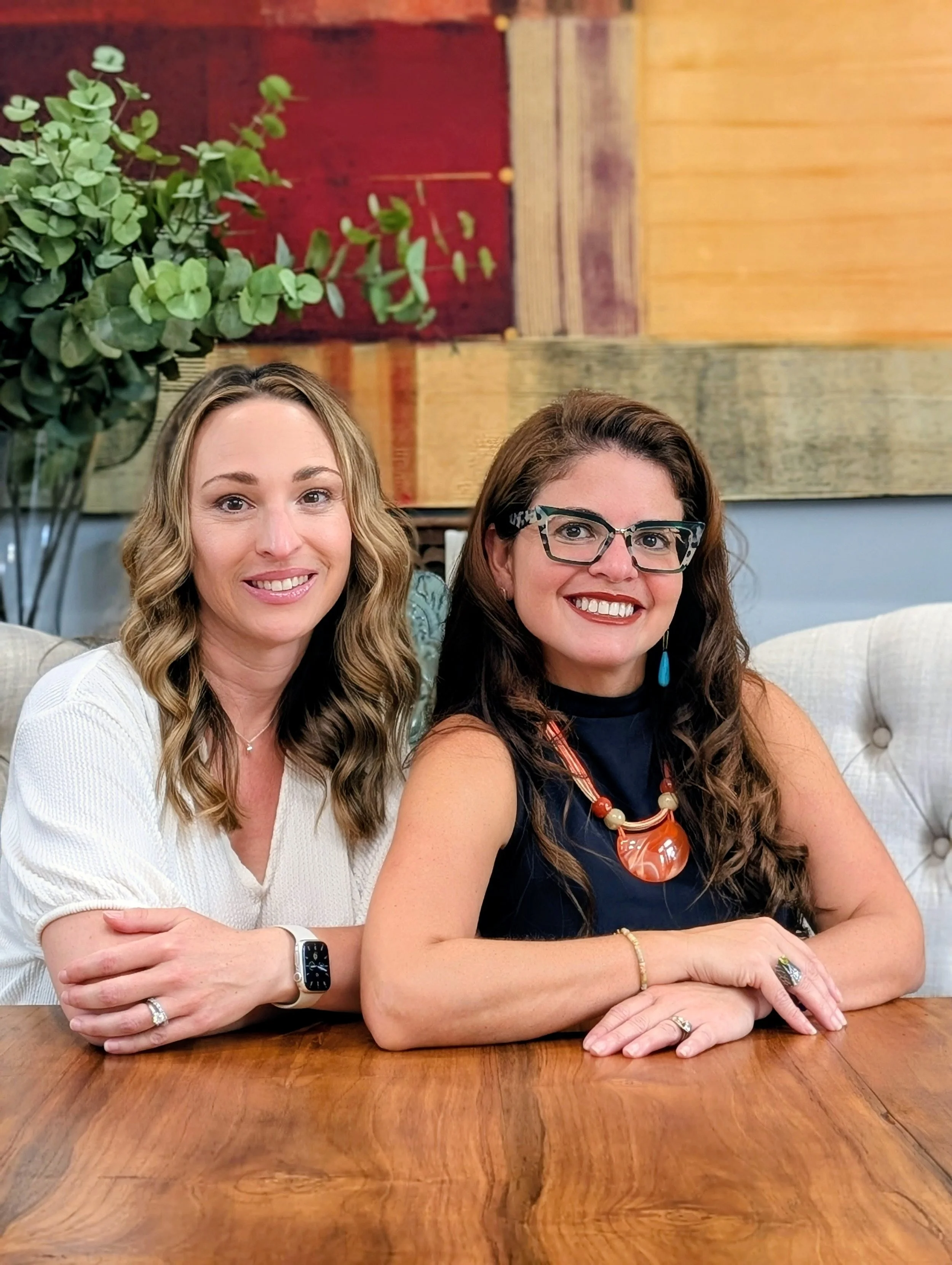 Two women sitting at a wooden table indoors, smiling at the camera. The woman on the left has wavy blonde hair, is wearing a white top and a smartwatch. The woman on the right has long curly brown hair, wears glasses, a dark sleeveless top, and colorful jewelry. There is a large green plant and abstract wall art in the background.