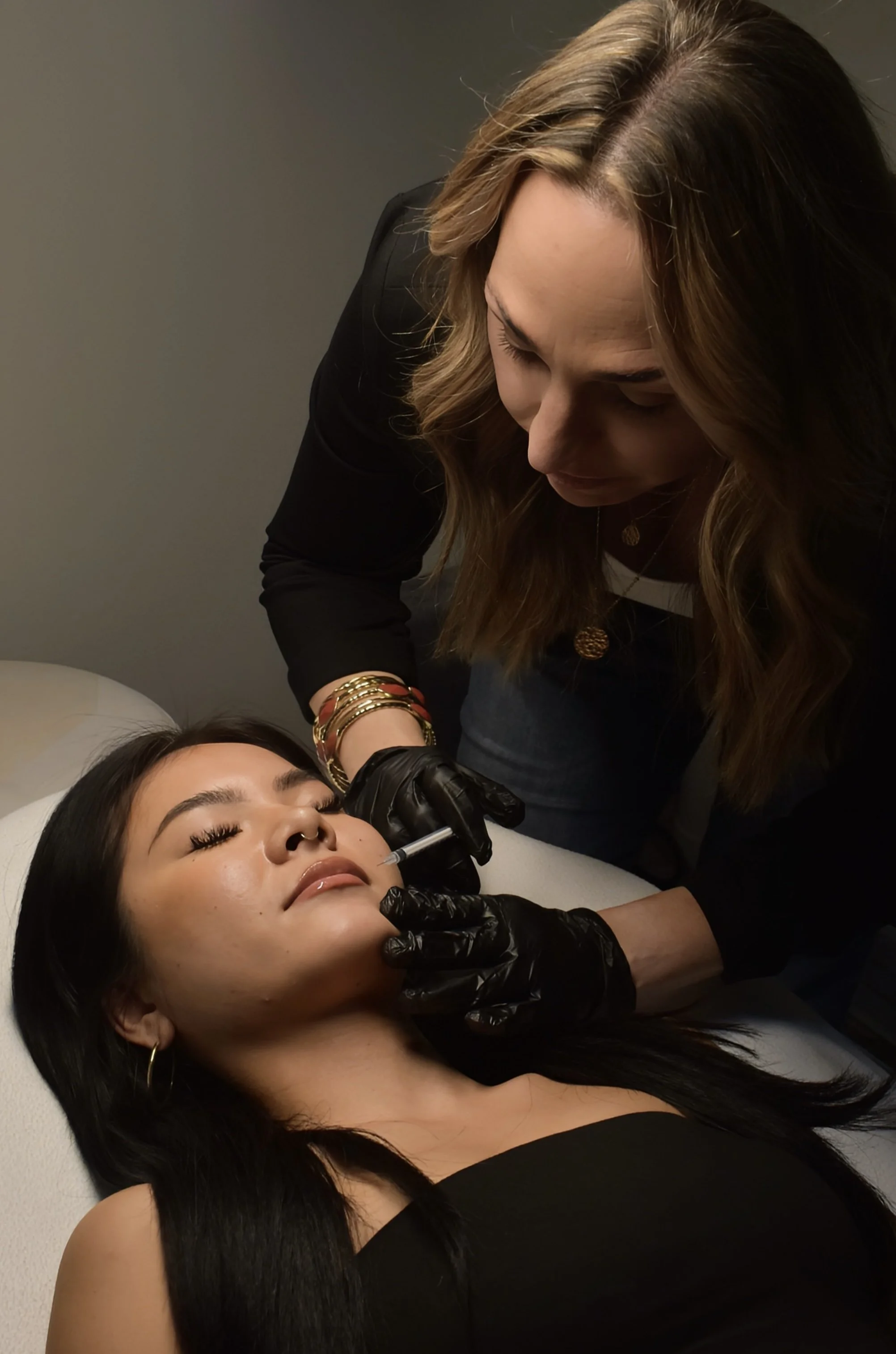 A woman laying on a medical bed receiving a cosmetic injection in her face from a professional wearing black gloves.