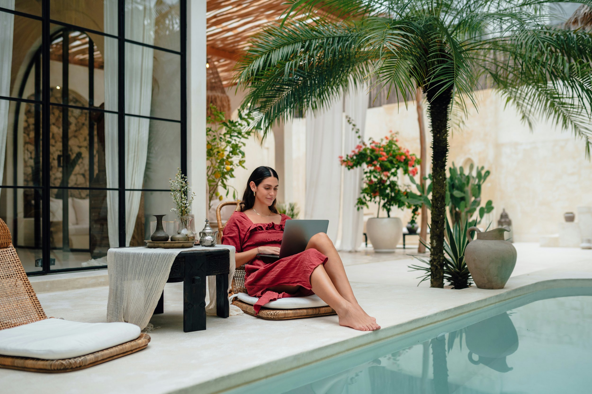 A young woman in a red dress sitting by a pool with her feet dipped in the water, working on a laptop. She is surrounded by lush indoor plants and decor, with white curtains and a large glass window in the background.