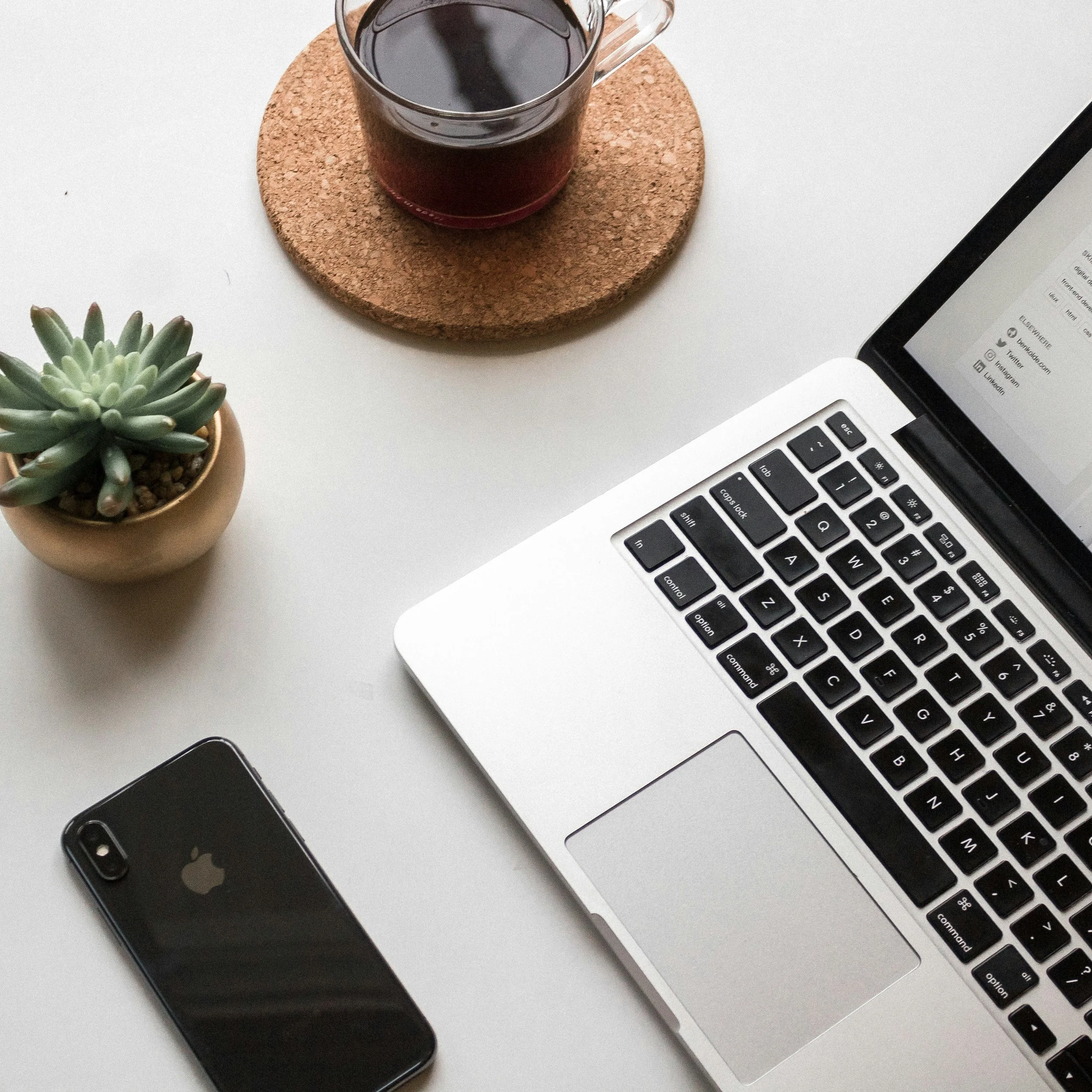A workspace with a MacBook laptop, an iPhone, a small potted succulent plant, and a cup of coffee on a cork coaster, all on a white surface.