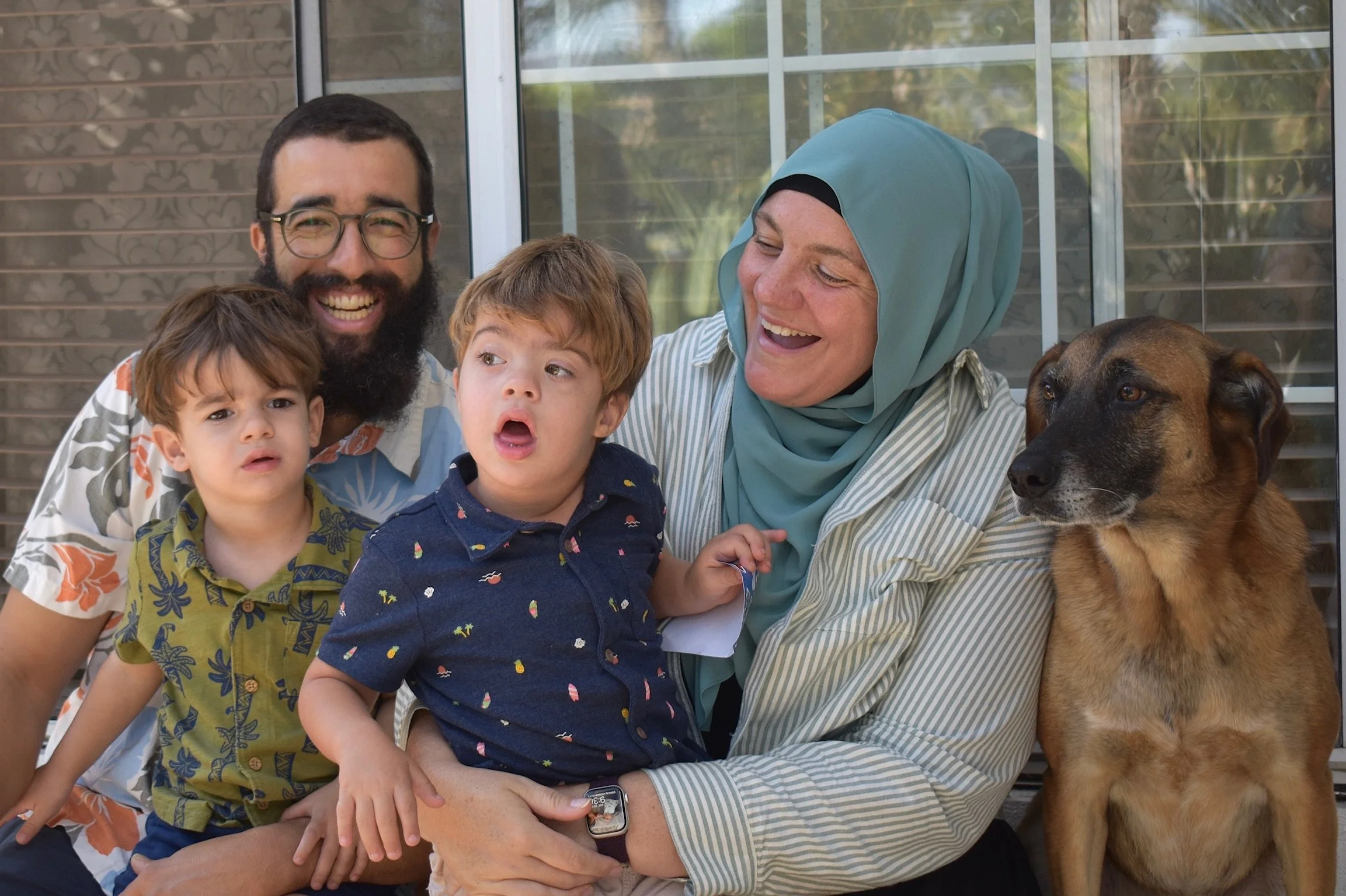 Family group photo on a porch with three children, two men, one woman with a headscarf, and a dog, all smiling and appearing happy.
