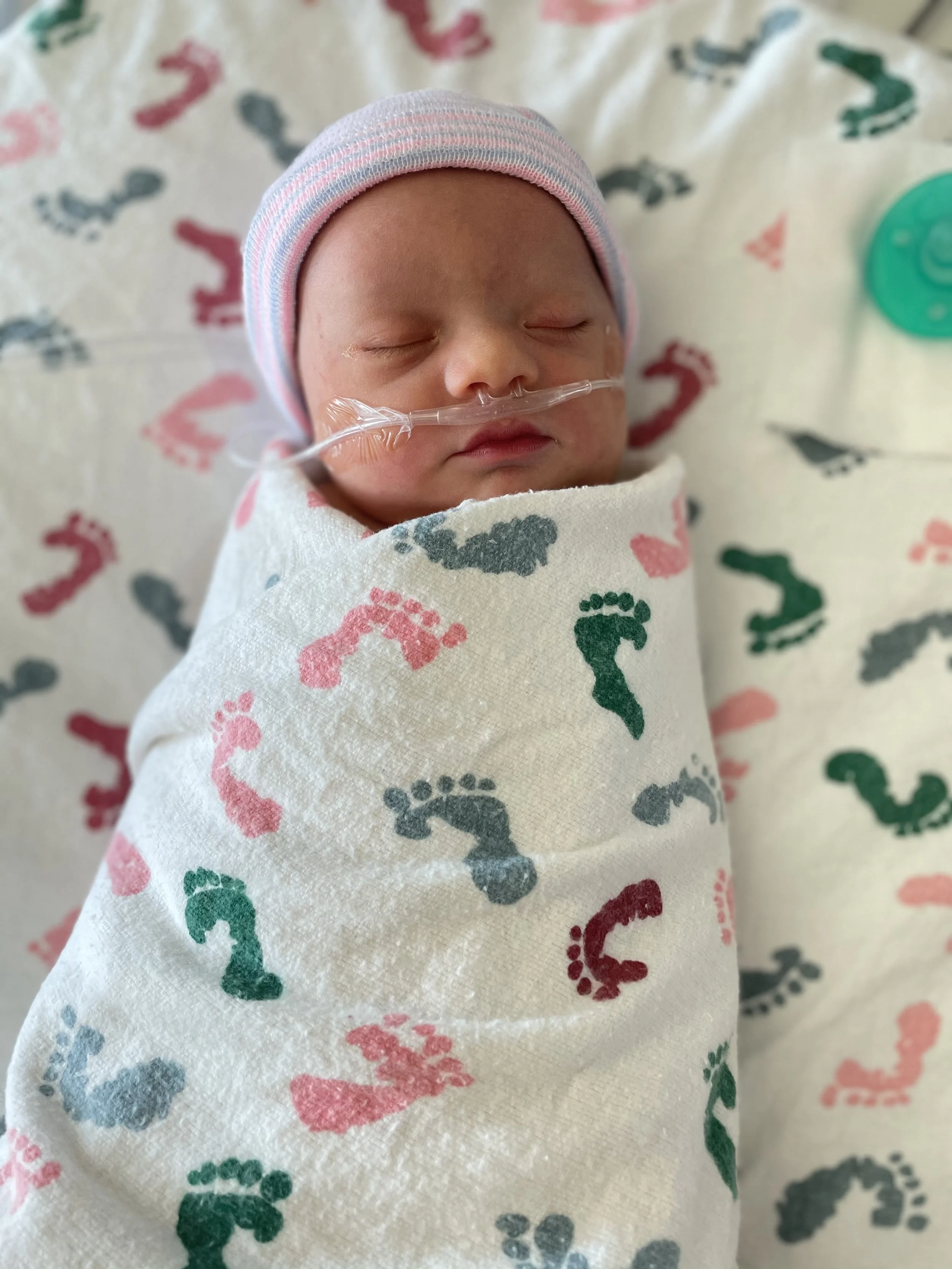 Newborn baby wrapped in a blanket with colorful footprints pattern, wearing a striped pink and blue hat, and with a nasal cannula for oxygen.
