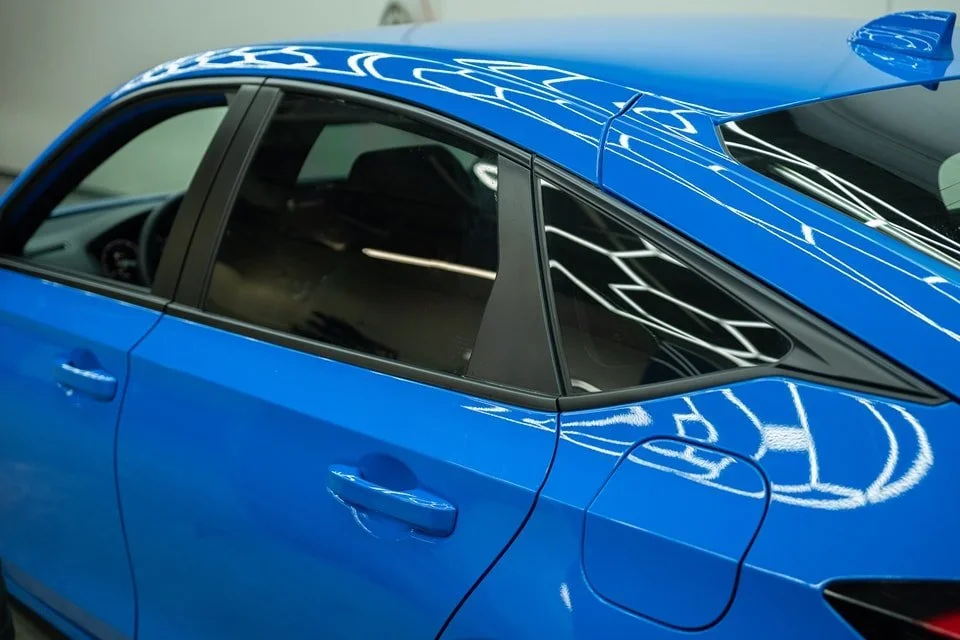 Close-up of a blue car showing the front passenger side, window, door handle, and part of the rear quarter panel with reflections of overhead lights.