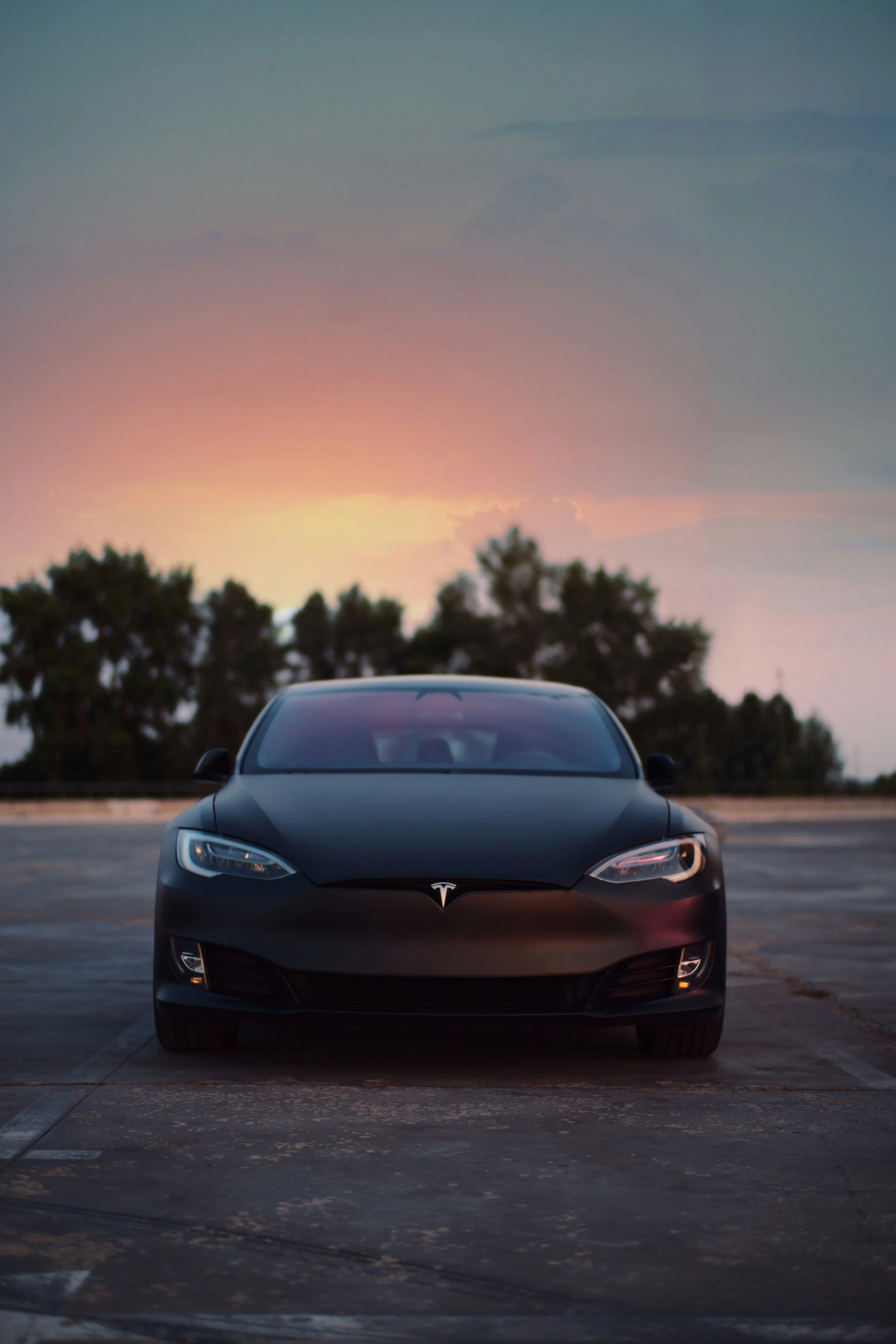 Black Tesla Model S parked on an empty lot at sunset with trees in the background.