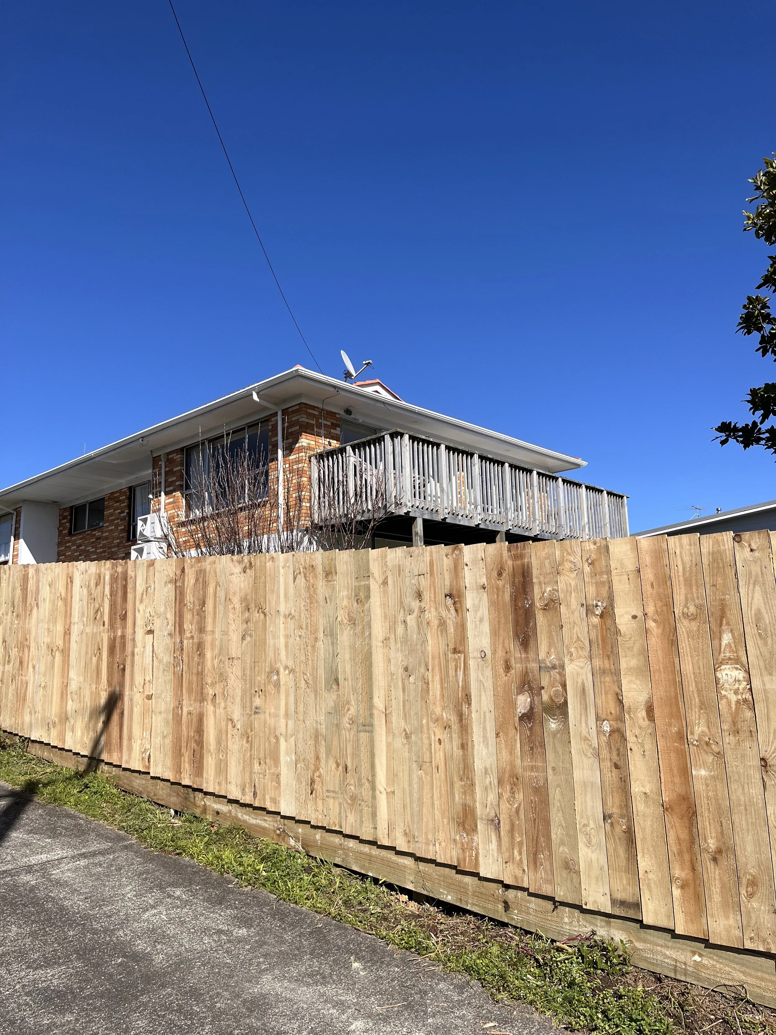 A two-story brick house with a wooden balcony on the second floor. There is a satellite dish on the roof and a wooden fence in the foreground. The sky is clear and blue.