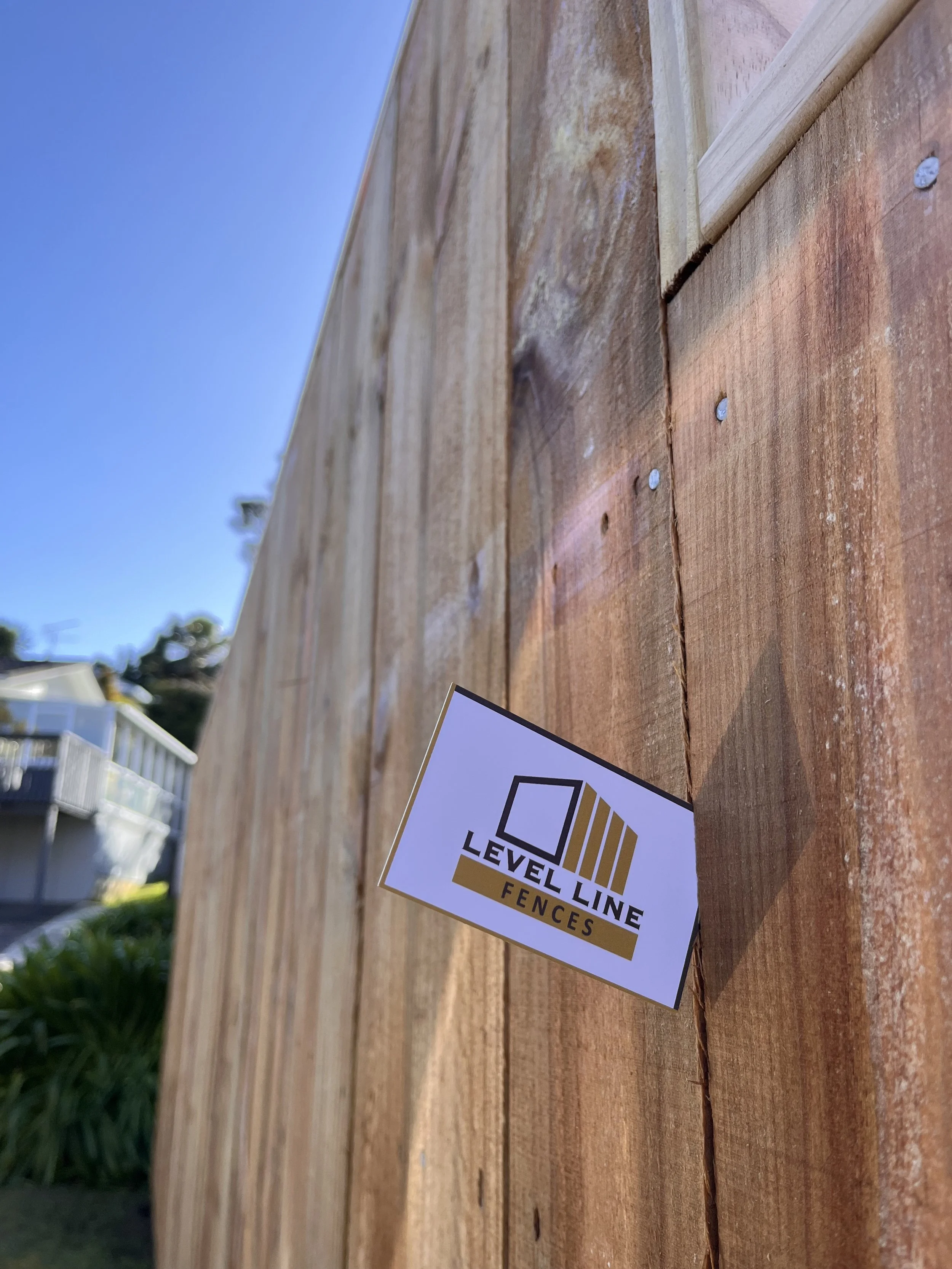 Close-up of a wooden fence with a Level Line Fences company sticker attached, showing a square logo and gold stripes, with a clear blue sky in the background.