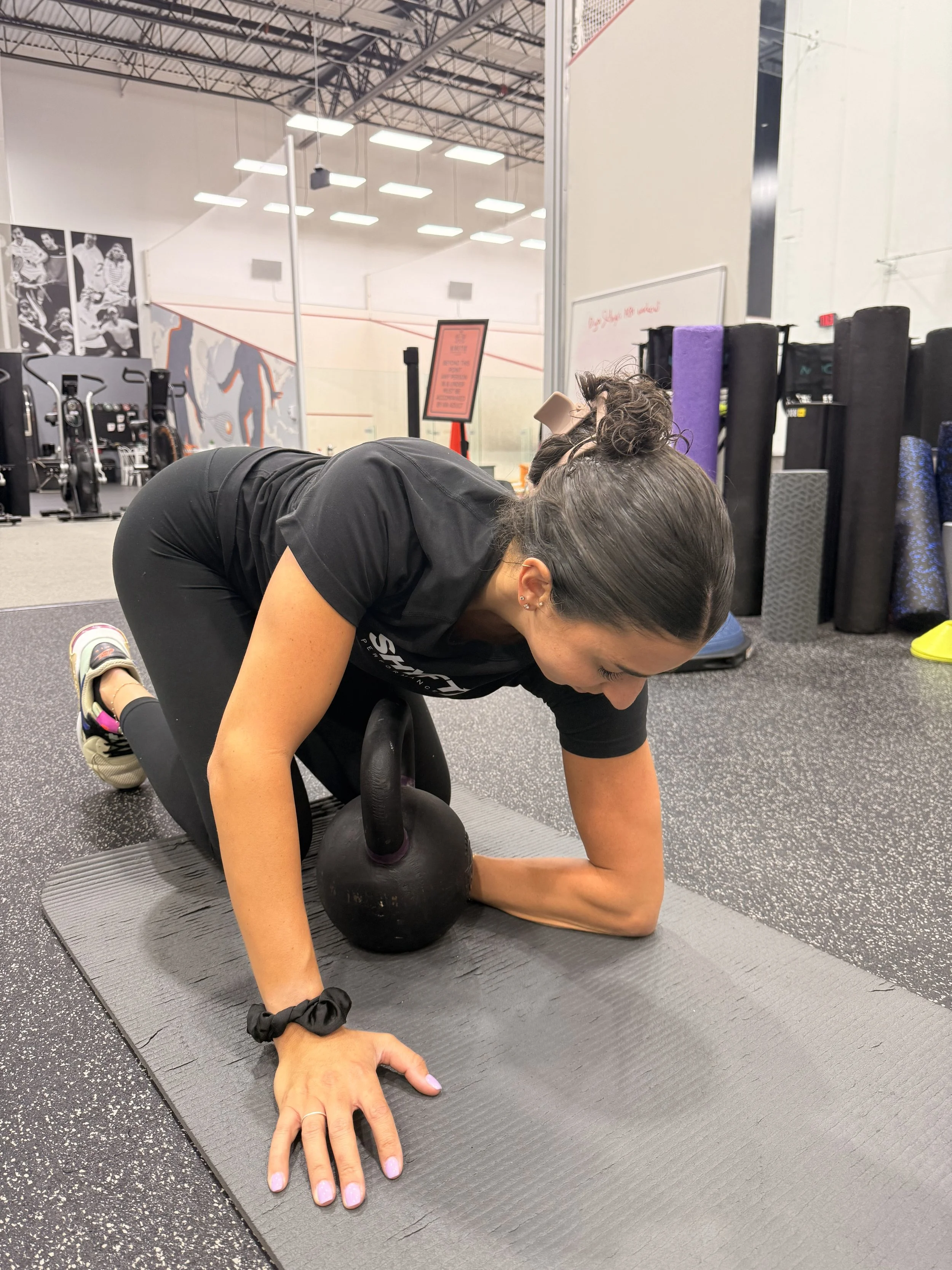 A woman is performing a kettlebell exercise on a mat in a gym. She is on her hands and knees with a kettlebell positioned near her torso. The gym has black flooring, fitness equipment, and foam rollers in the background.