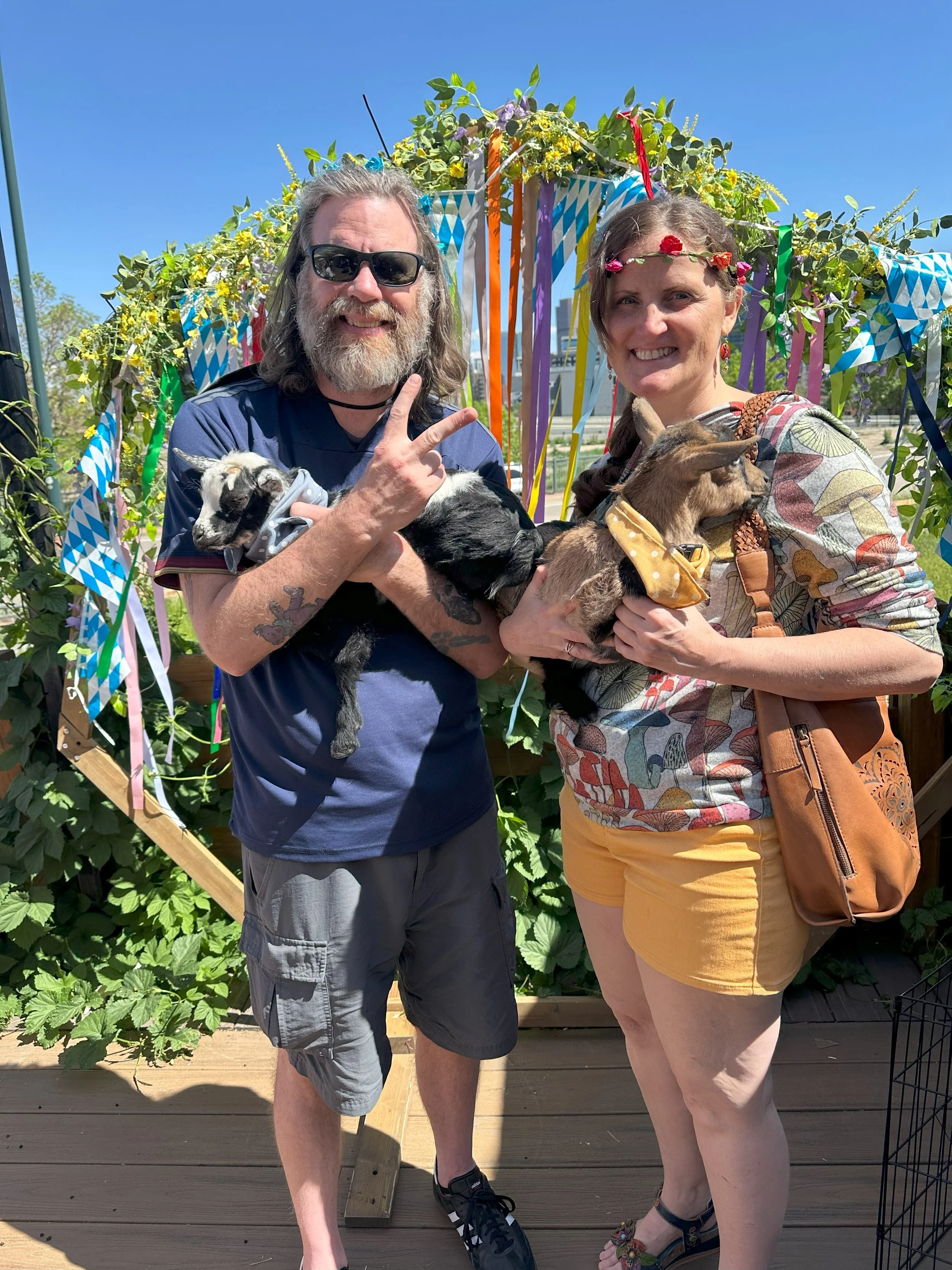 A man and woman stand outdoors on a sunny day, holding puppies, with a decorative backdrop covered in greenery and colorful ribbons.
