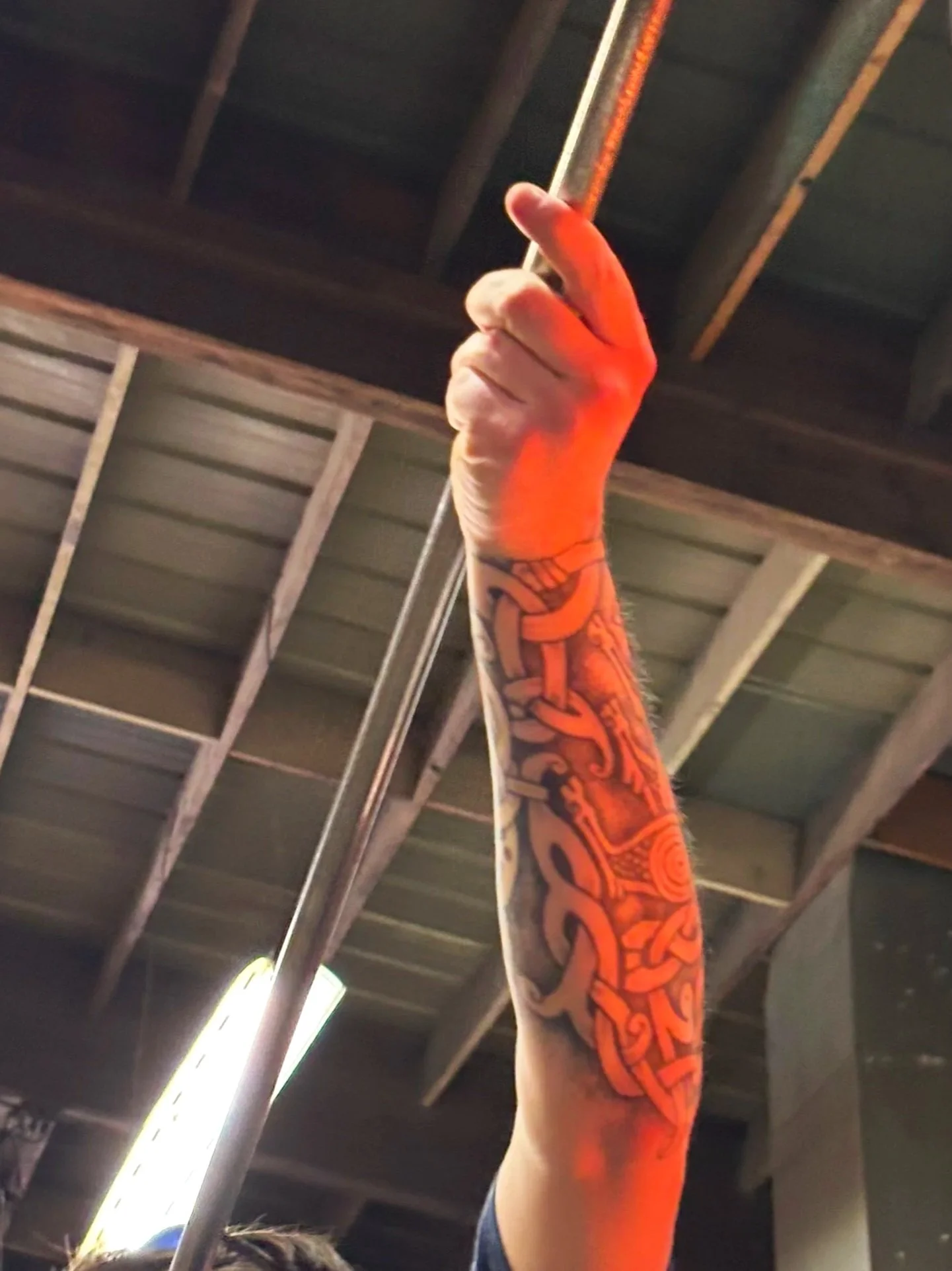 A person with a colorful tattoo sleeve on their arm holding a metal pole in a gym or warehouse setting with industrial ceiling beams.