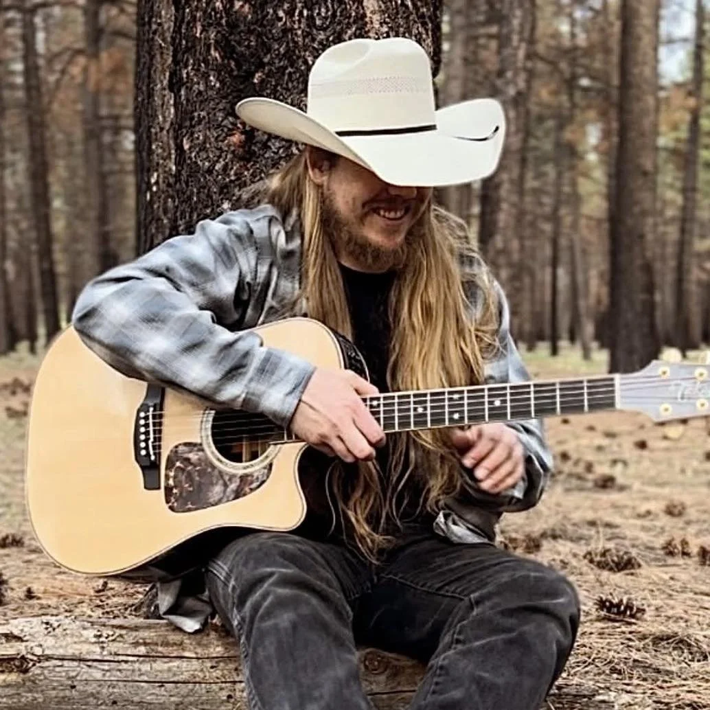 A man with long hair and a beard, wearing a cowboy hat and a plaid shirt, sitting in a forest and playing an acoustic guitar.