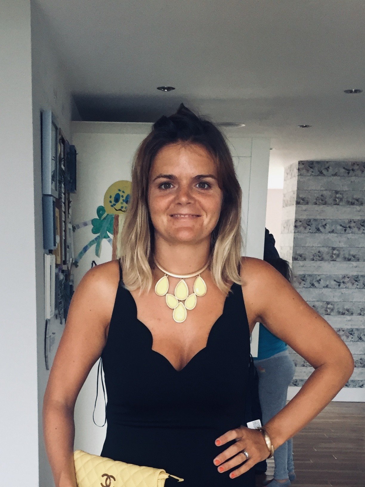 A woman with shoulder-length blonde hair wearing a black dress and a beige statement necklace, holding a yellow quilted purse with a Chanel logo, standing indoors against a light-colored wall with some artwork and a stone feature wall in the background.