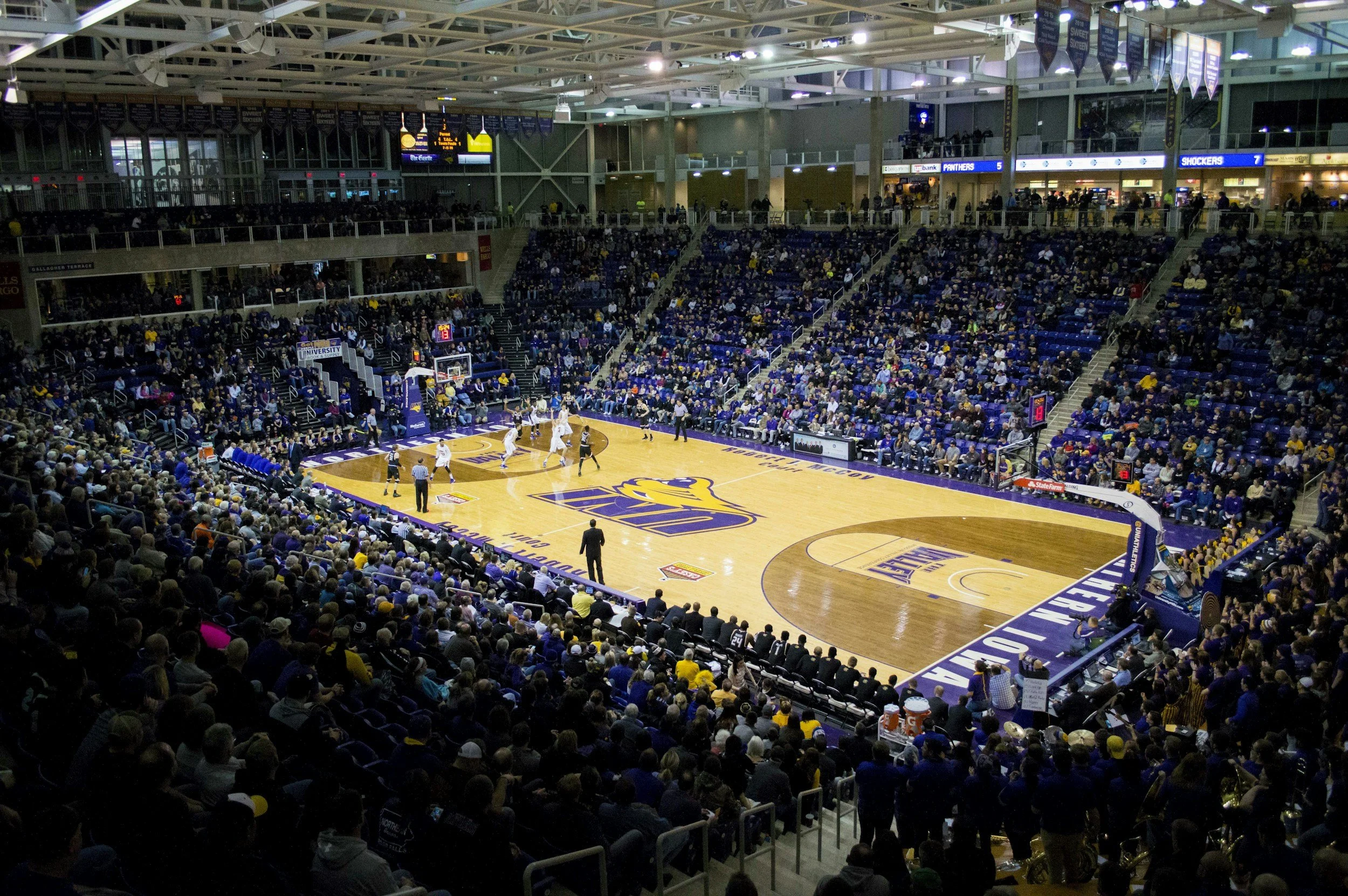 An indoor basketball game at the University of Northern Colorado's Greeley arena, with players on the court, referees, and a full crowd of spectators.