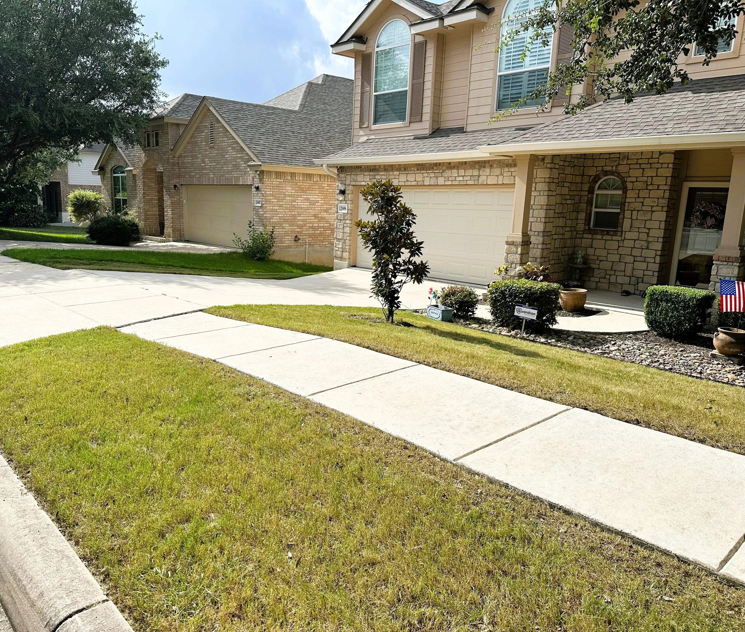 Front view of a house with a brick and siding exterior, and a driveway with a sidewalk leading to the entrance. There are bushes, potted plants, and an American flag beside the house.