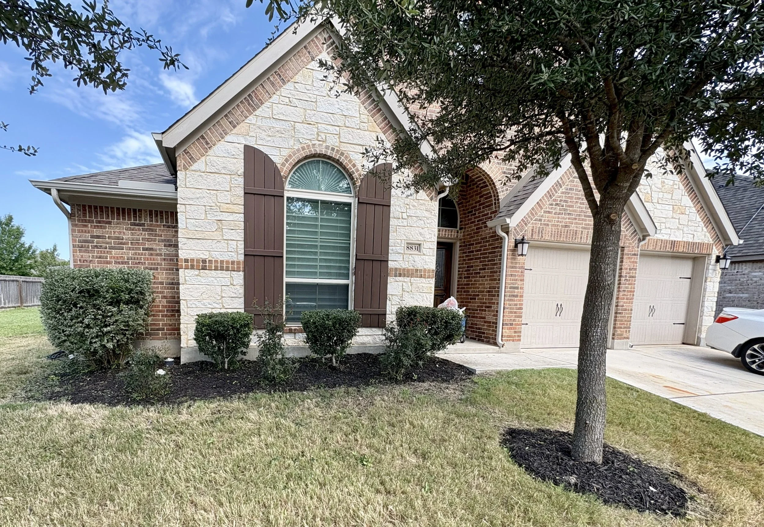Front view of a brick and stone house with a two-car garage, a sidewalk, a tree, and a small front yard with bushes and grass.