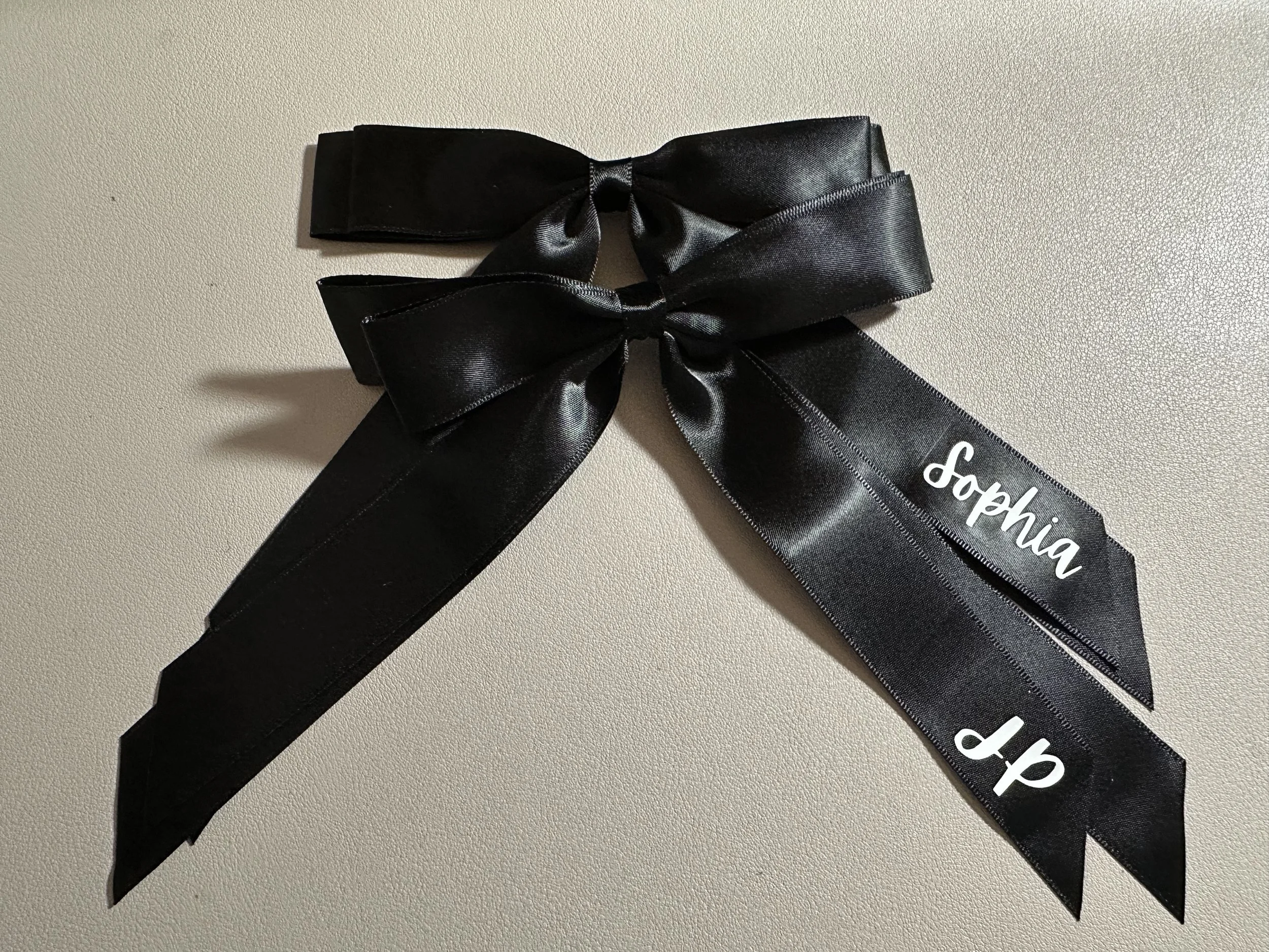 Black satin ribbon bow with the name Sophia embroidered in white on one of the tails.
