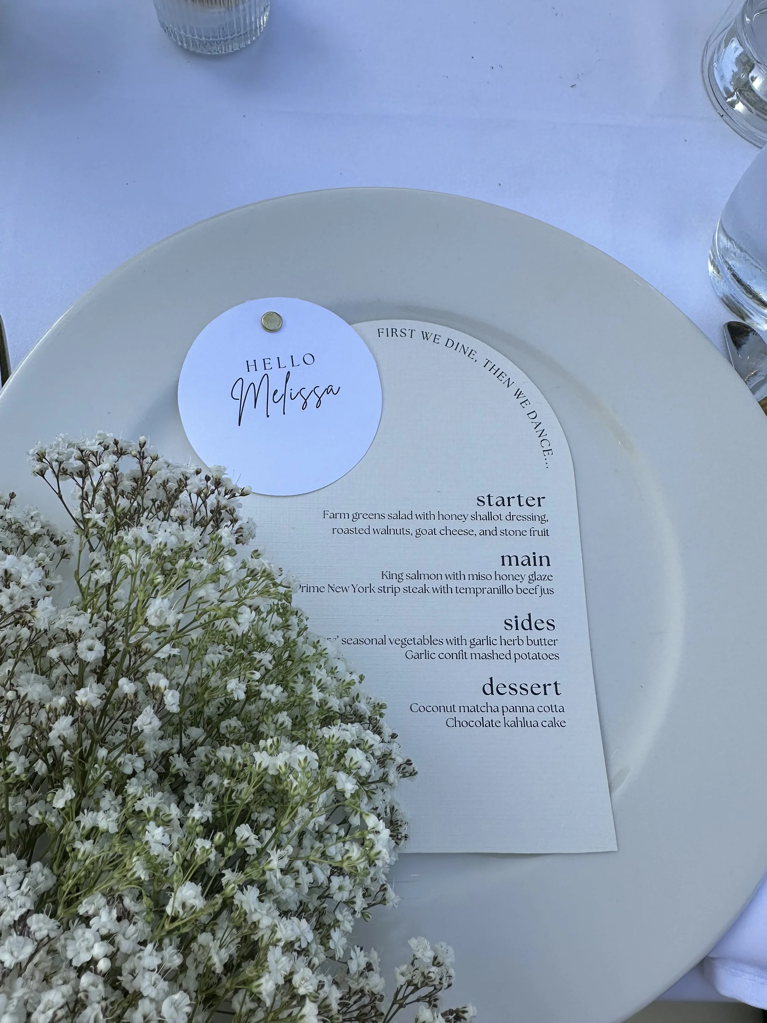 A table setting with a white plate, a floral arrangement of small white flowers, and a dinner menu with a personalized name tag reading 'Hello Melissa'. The menu lists a four-course meal including starter, main, sides, and dessert, and features a drink at the top of the menu.
