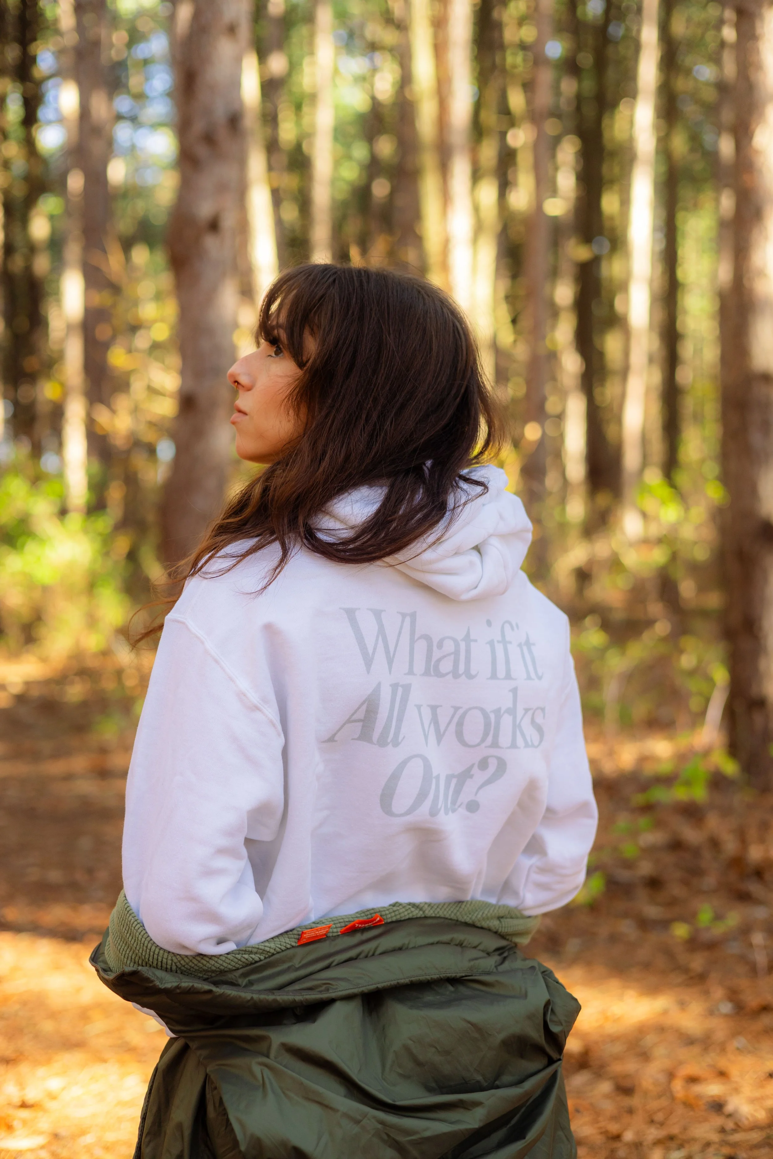 A woman is standing in a forest with tall trees and sunlight filtering through the leaves. She is wearing a white hoodie with text on the back that reads, 'What if all words out?'. She has dark brown, wavy hair and is turned slightly to the side, gazing into the distance.