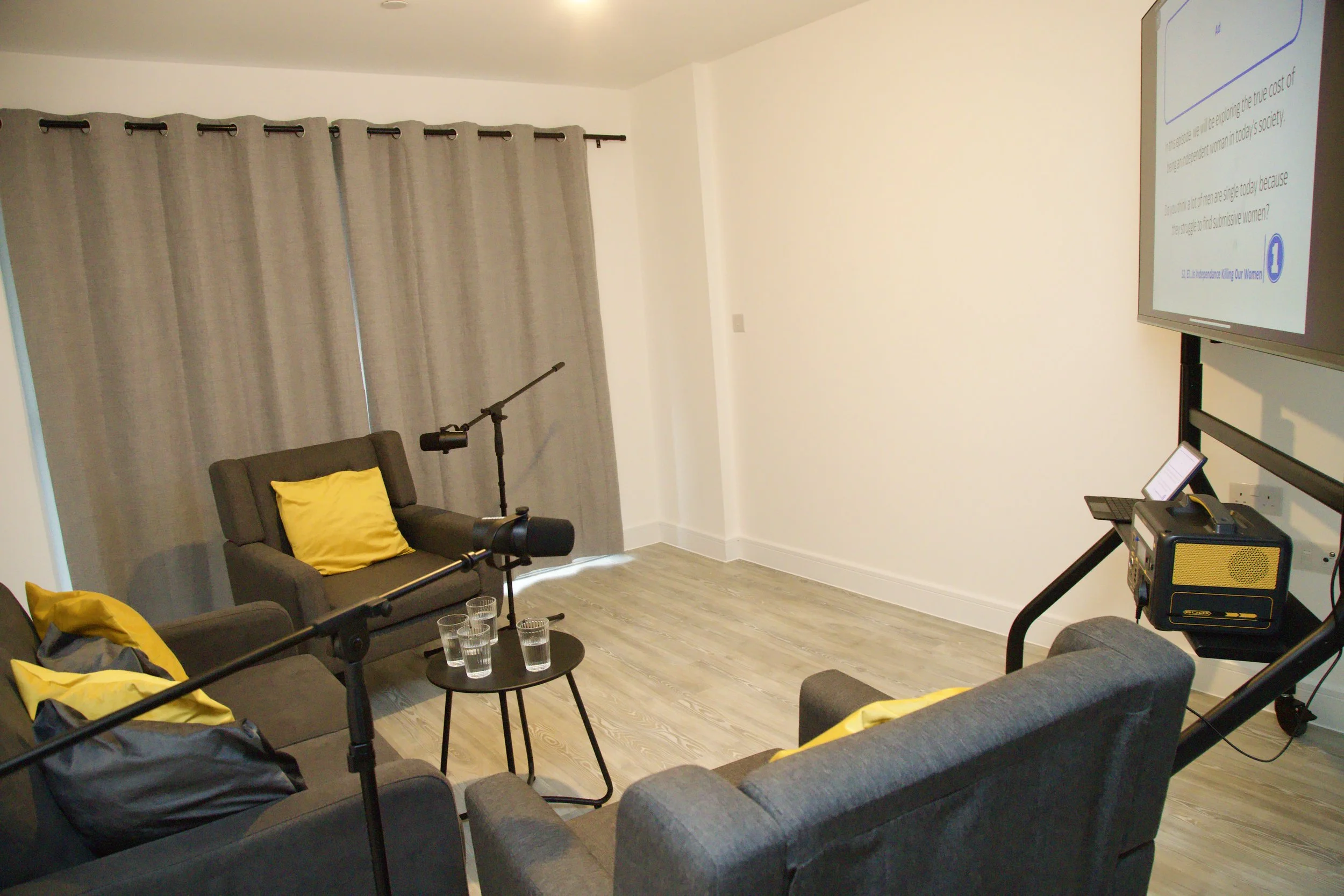 A modern living room with two gray couches, yellow pillows, a small black table with four glasses, and a large TV screen displaying a presentation. There is a microphone on a stand in front of one of the couches, and gray curtains covering a window o