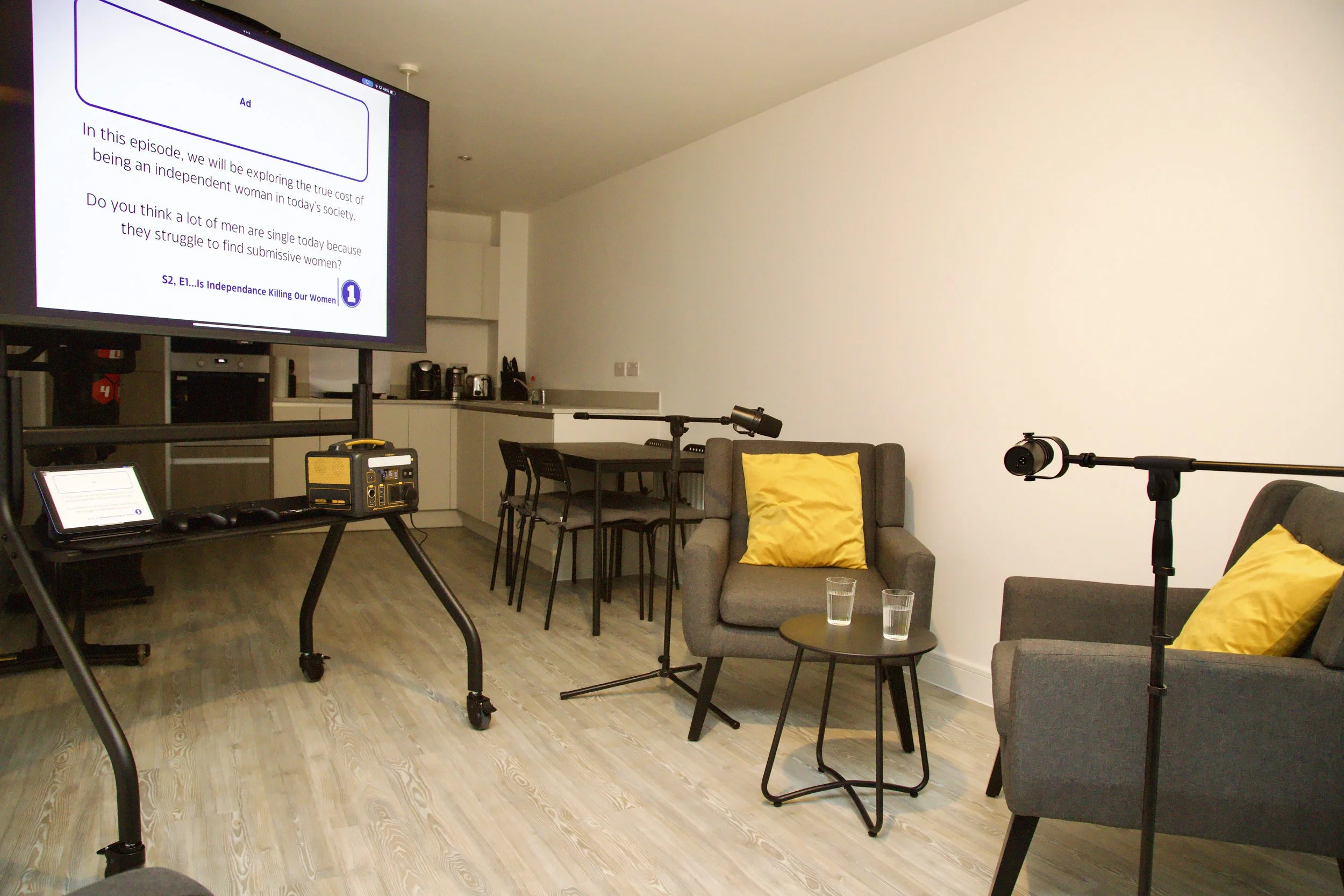 Living room setup with a large TV, a tablet, a yellow toolbox, two gray armchairs with yellow pillows, a small round table with two glasses, and audio/recording equipment including a microphone and boom arm, with a kitchen and dining area in the back