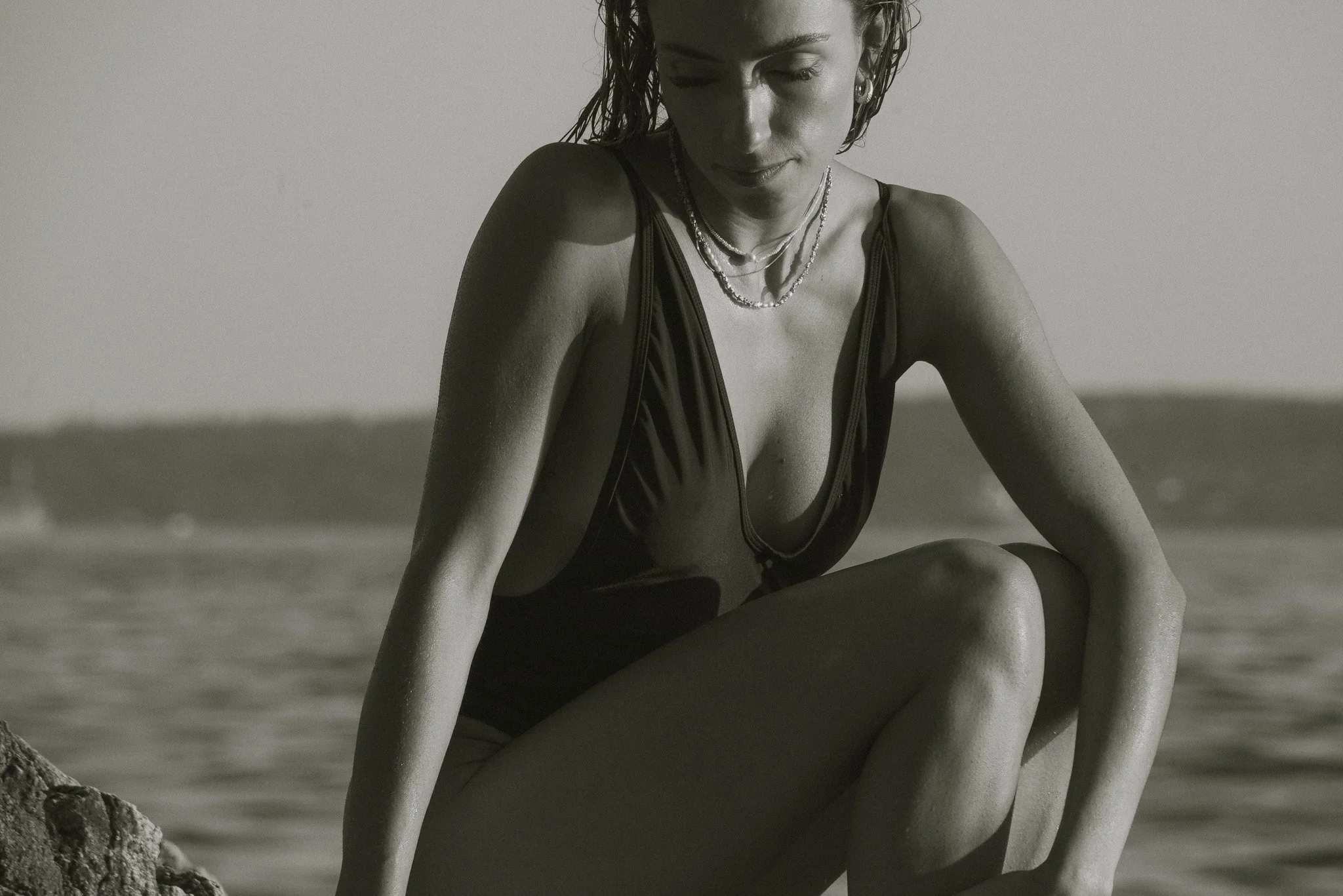 A woman sitting on the beach, wearing a swimsuit and layered necklaces, with wet hair, looking down with a serene expression.