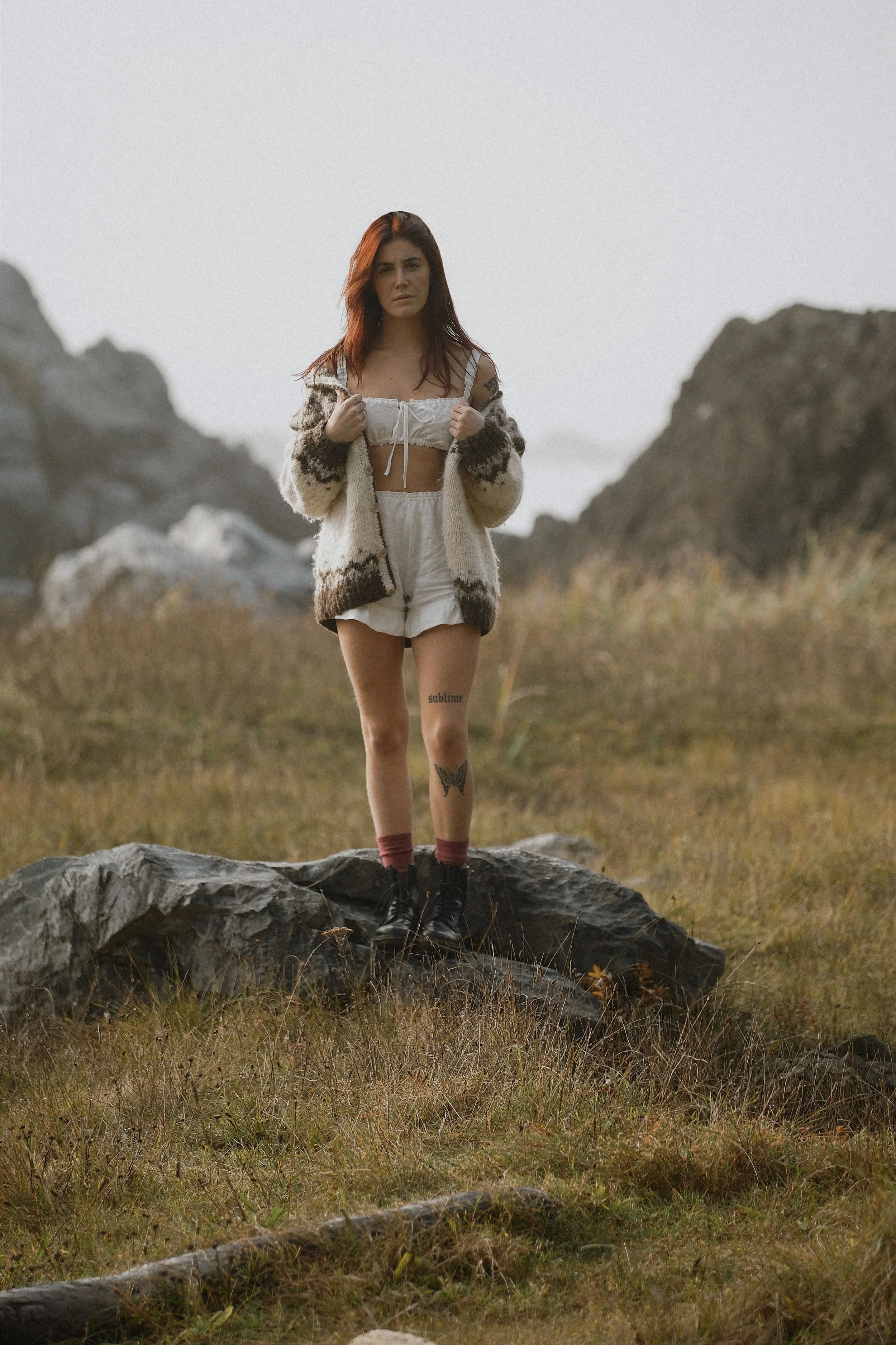 A young woman with red hair stands on a large rock in an outdoor natural setting, surrounded by grass and rocks, with mountains in the background. She is wearing a cozy, patterned cardigan over a crop top and shorts and has visible tattoos on her left thigh and right arm.