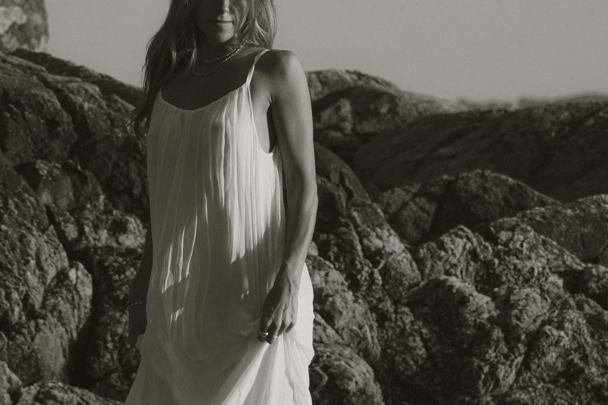 A woman in a flowing white dress with spaghetti straps standing on rocks, looking to the side with waves in the background.
