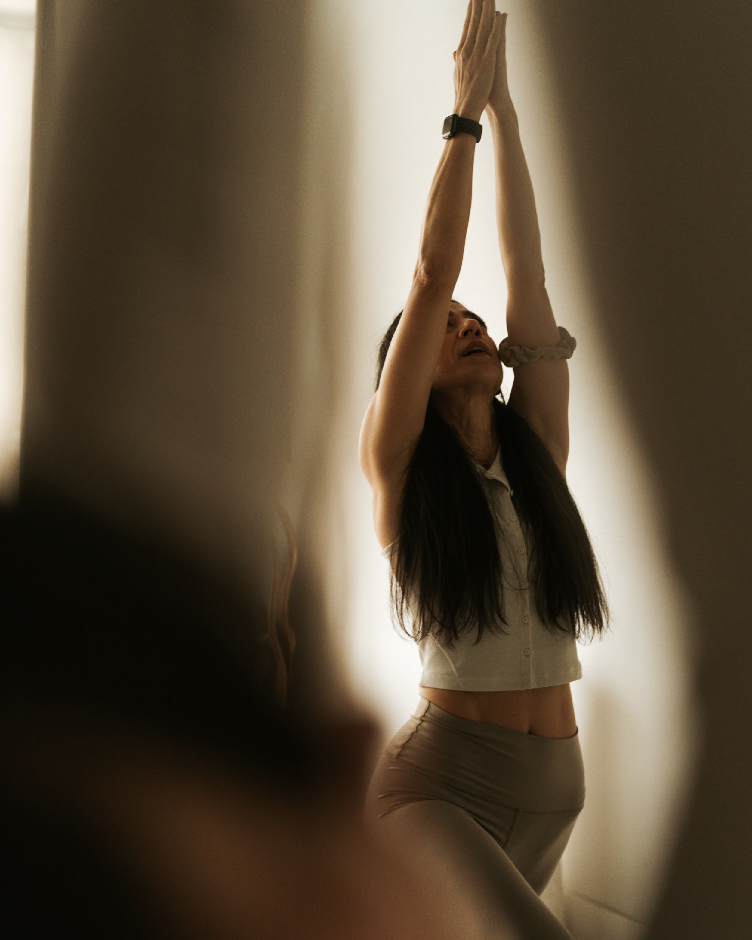 A woman with long dark hair is standing with her arms raised above her head, hands pressed together, in a pose in front of a bright, softly-lit background. She is wearing a fitted beige tank top and beige pants, with a watch on her wrist.