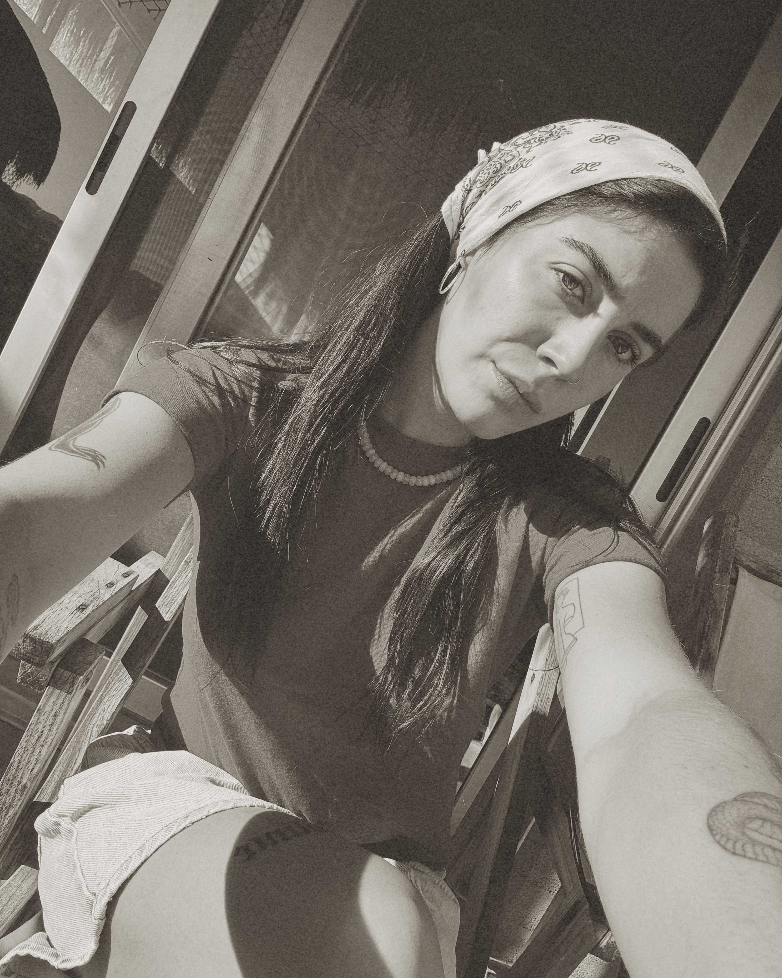 A person with long hair, wearing a bandana, a necklace, and earrings, looking at the camera while taking a selfie in front of an elevator with metallic doors, and part of a person's arm and foot is visible in the foreground.