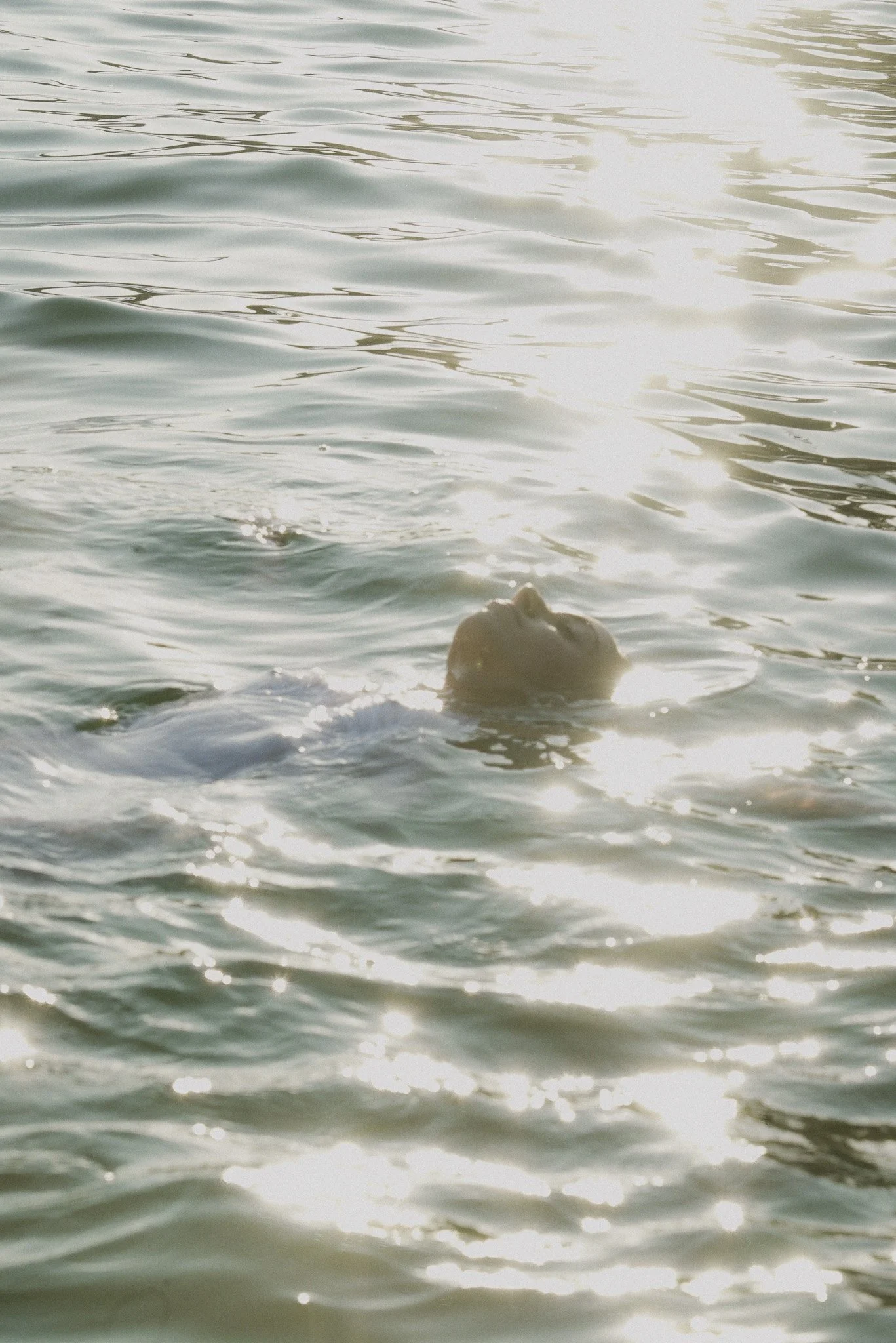 A person floating in a body of water with their face partially submerged, with sunlight reflecting off the water surface.