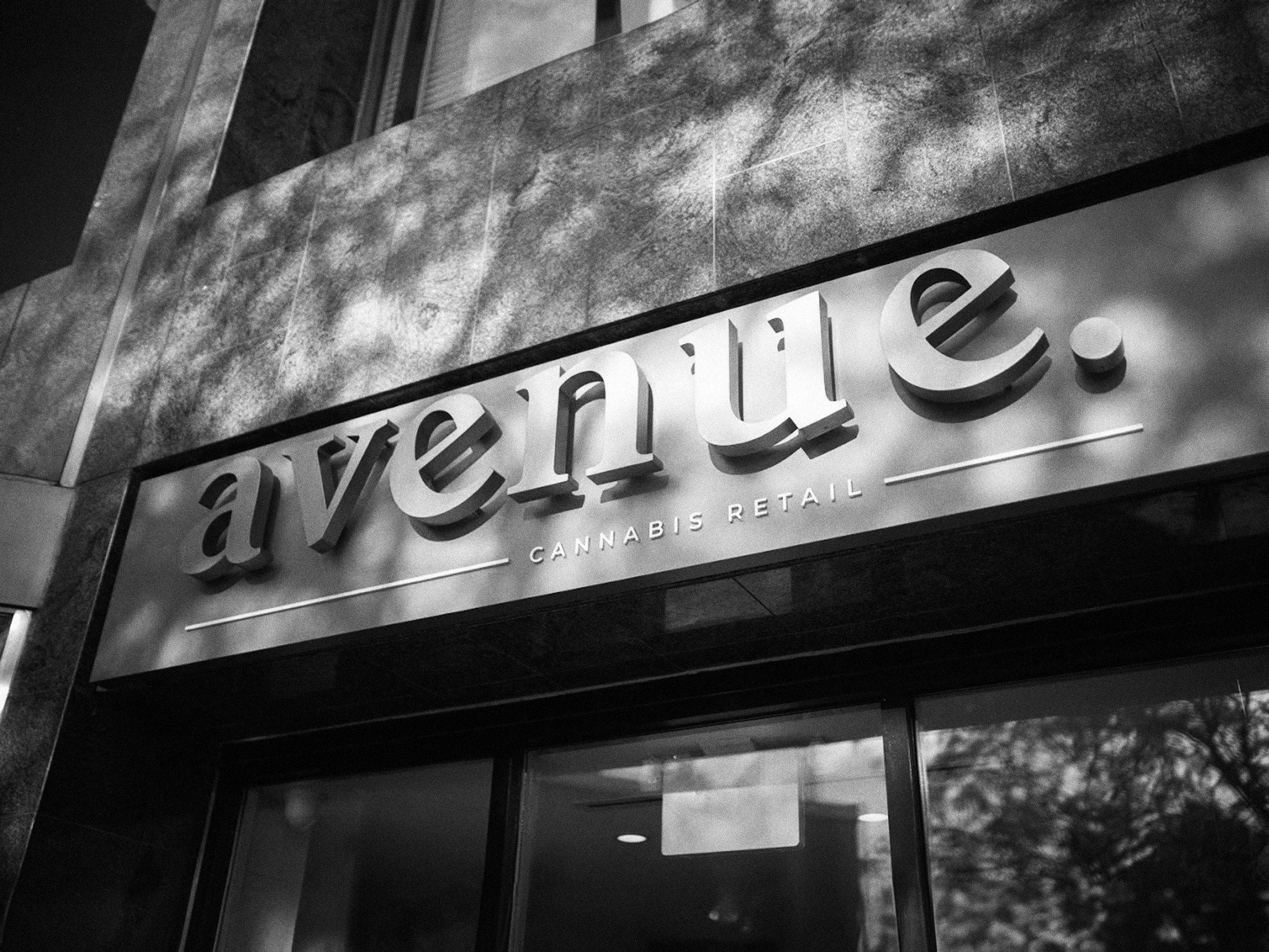 Close-up of a storefront sign reading 'ave.pl' with part of the building's exterior wall above, in black and white.