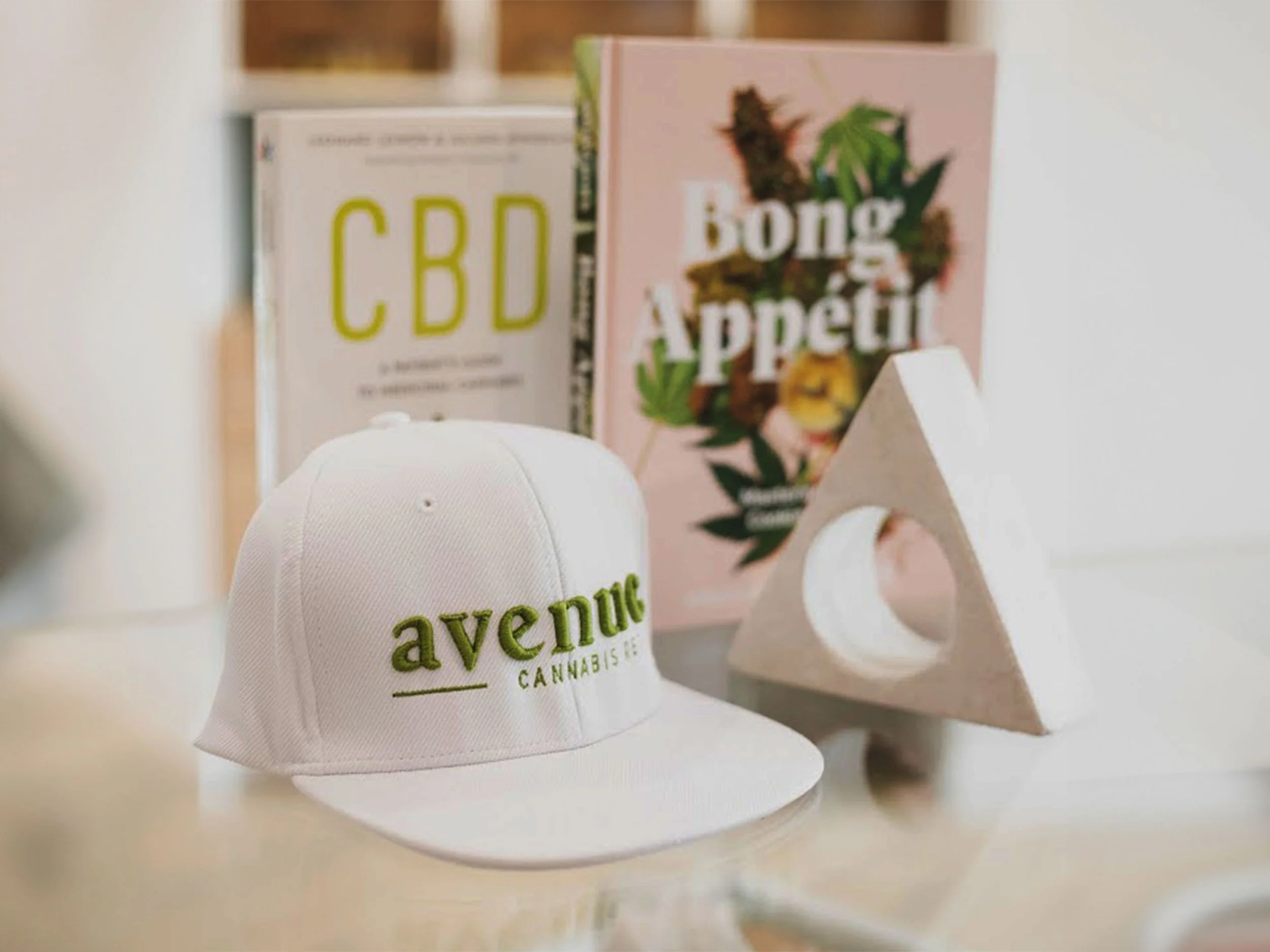 A white baseball cap with the words "avenue CANNABIS" embroidered in green on the front, placed on a white surface. In the background, there are books and a decorative plant.