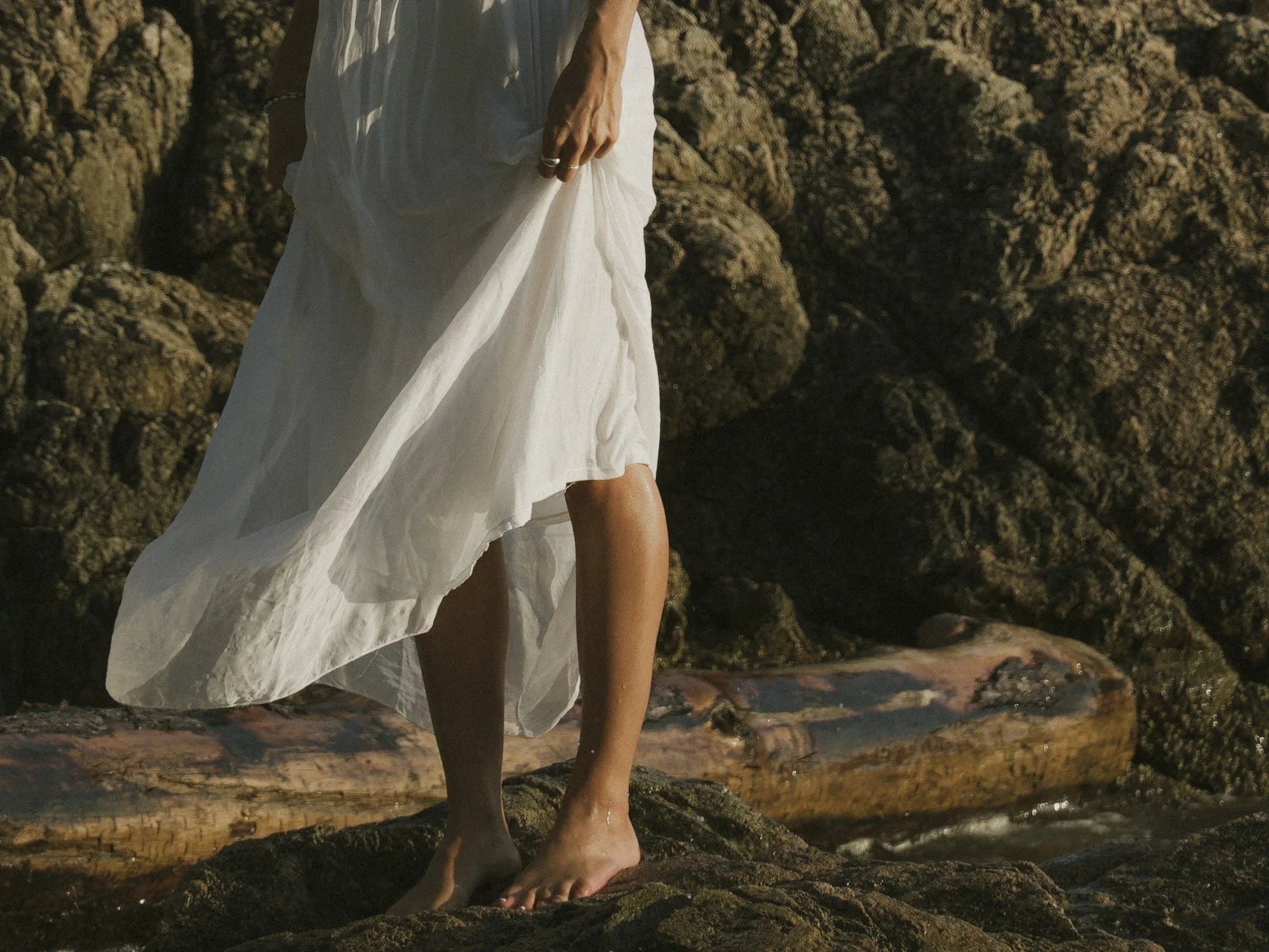 A person in a white dress standing barefoot on a rocky beach with the ocean in the background.