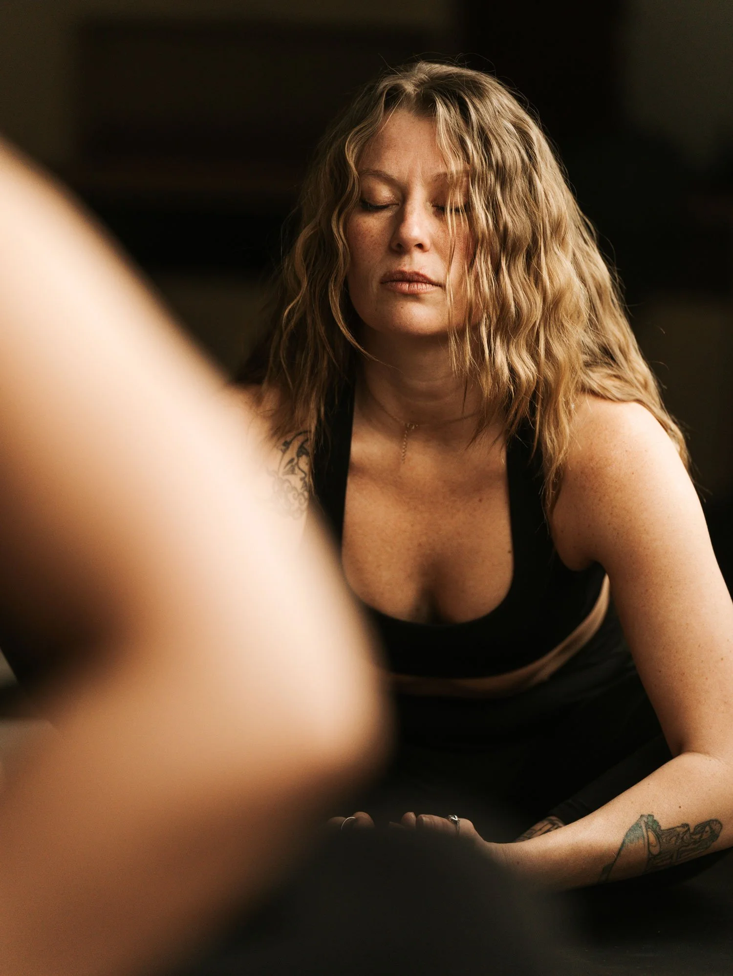 A woman with curly hair and tattoos on her arm, eyes closed, in a black top, with a dark background.
