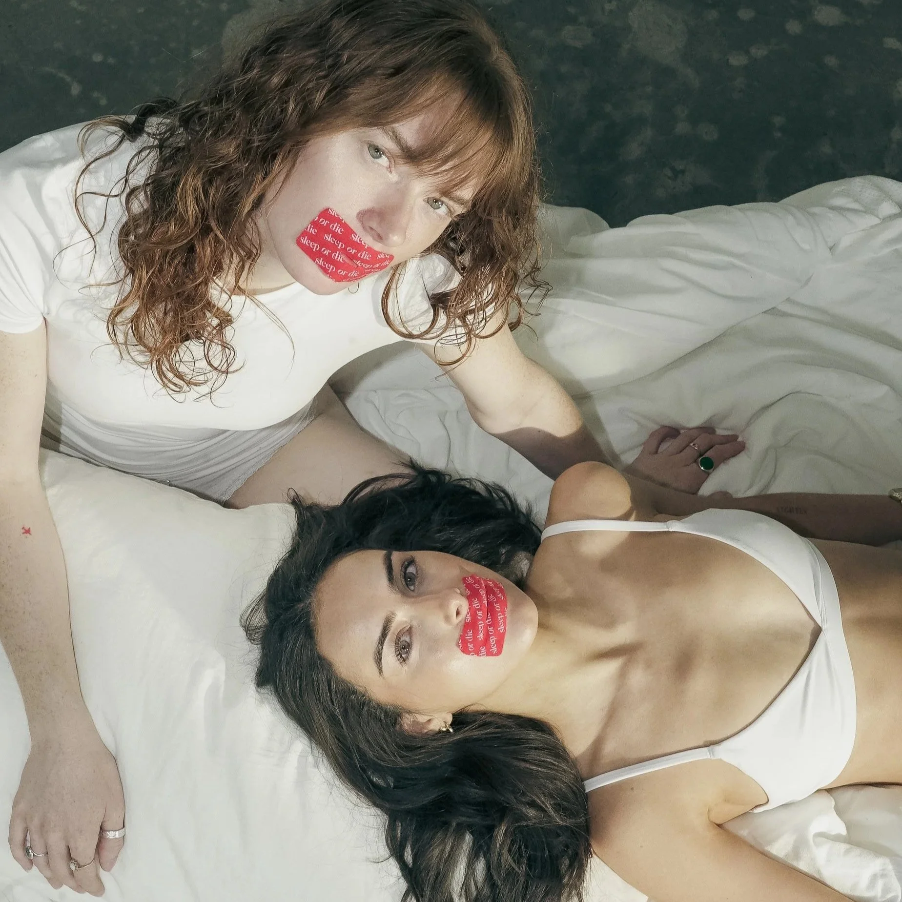 Two women lying on a white bed, one with curly red hair and the other with dark hair. Both women have red stickers over their mouths that say 'sleep or die.' The woman with dark hair is wearing a white tank top.