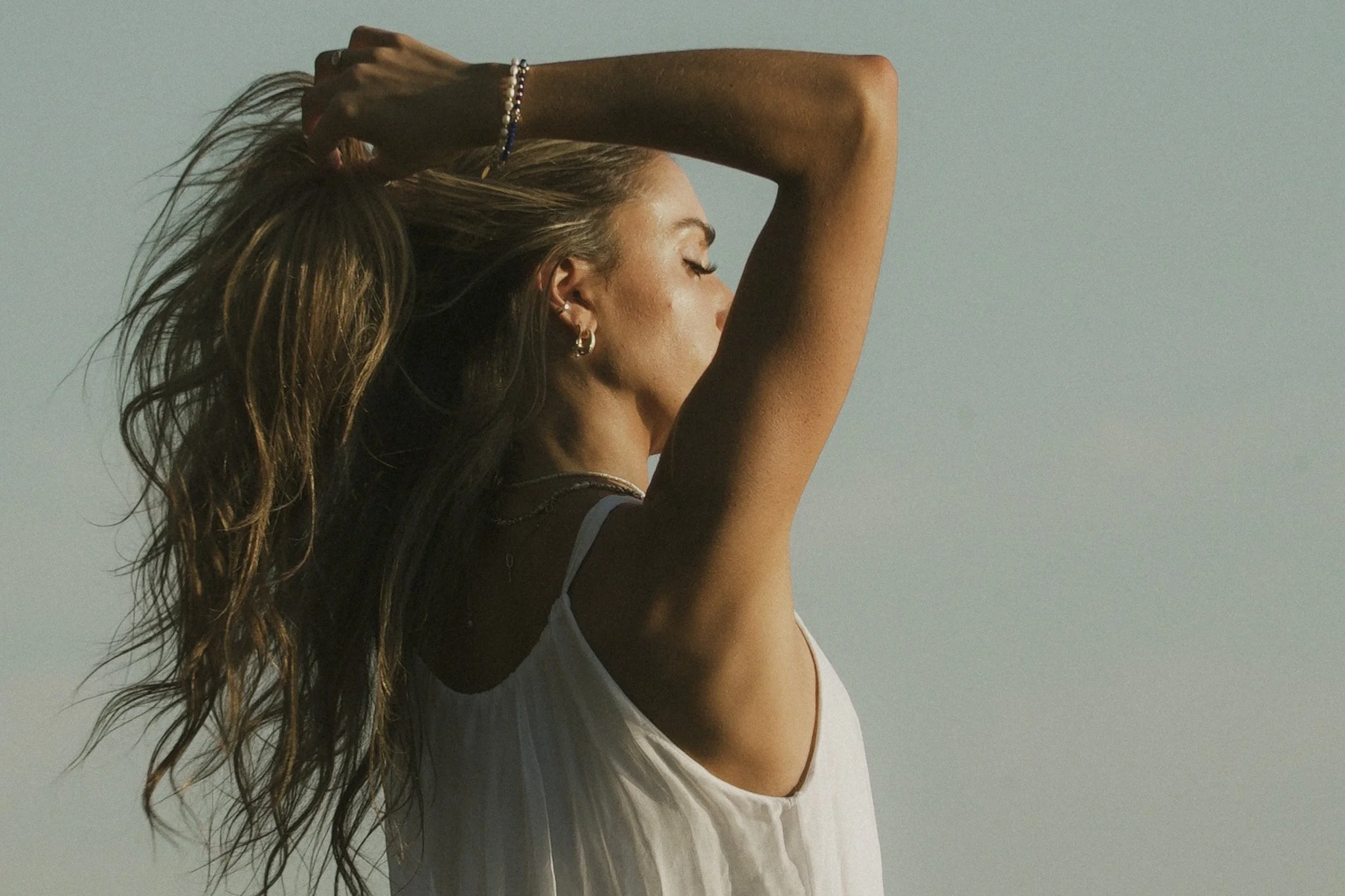 A woman with long wavy hair and multiple earrings, wearing a white sleeveless top, is standing against a plain sky, with her eyes closed and her hand resting on her head.
