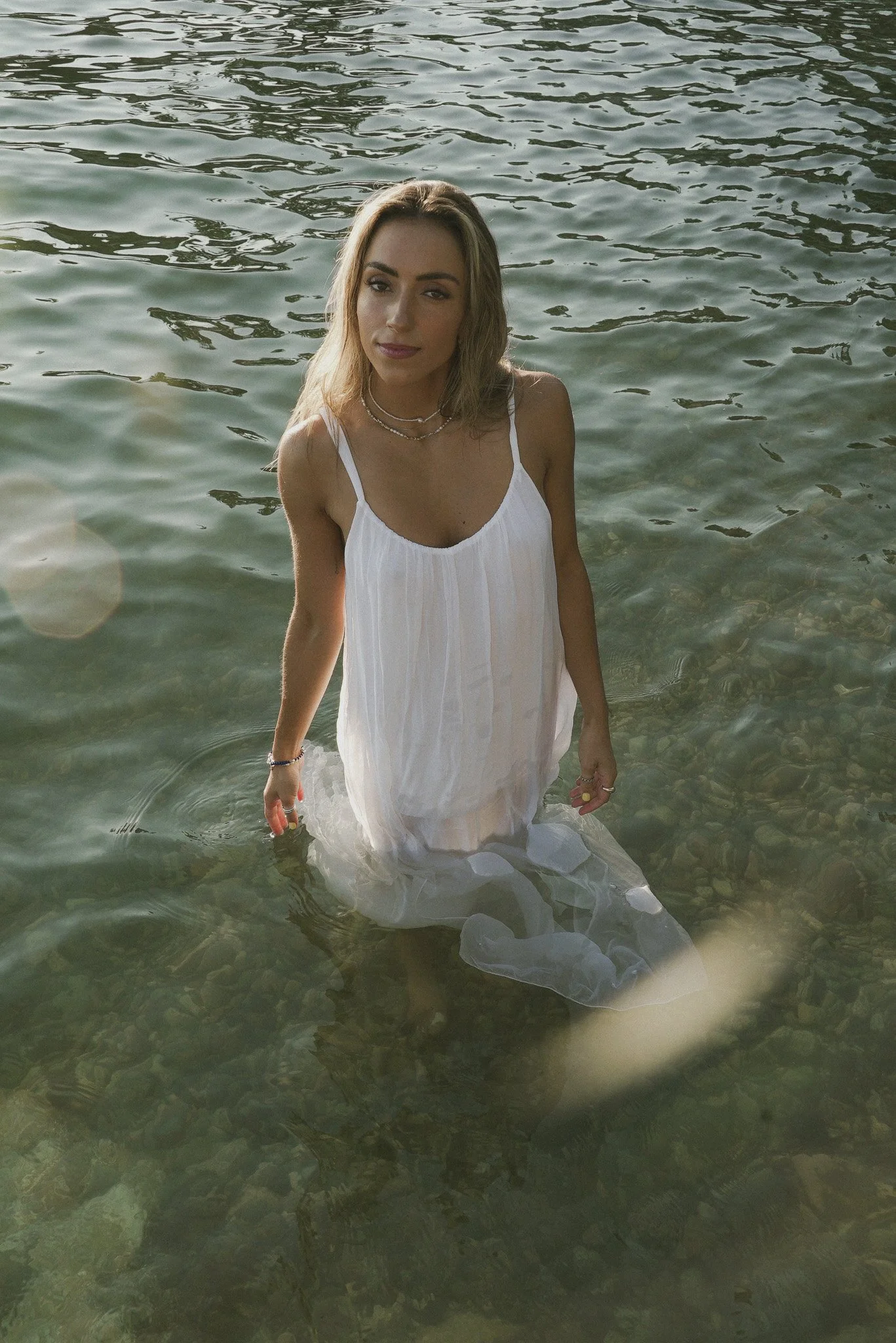A woman standing in clear water, wearing a white dress with spaghetti straps, looking at the camera with a slight smile. The water reaches just below her waist, and the background shows rippling water with some reflections.