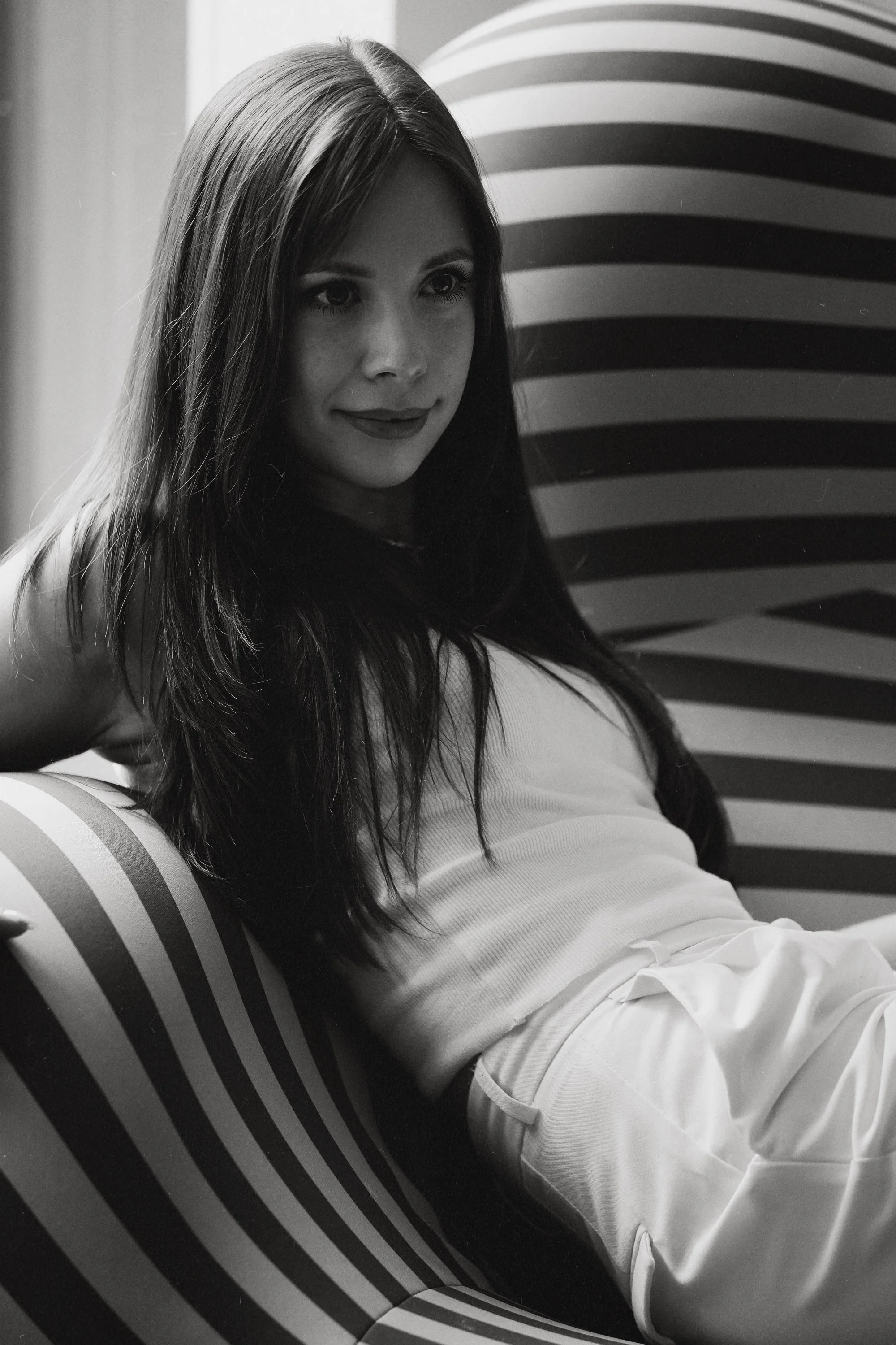 A woman with long dark hair sitting on a striped cushioned chair, wearing a sleeveless top and light-colored pants, looking to her right in a black and white photo.