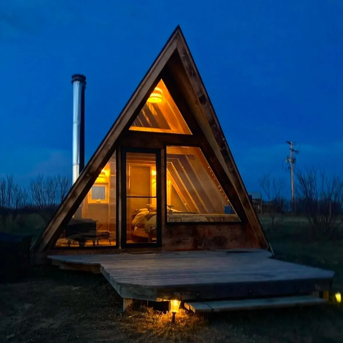 A small A-frame cabin with warm interior lighting, a wooden deck, and a chimney, set against a dark blue evening sky.