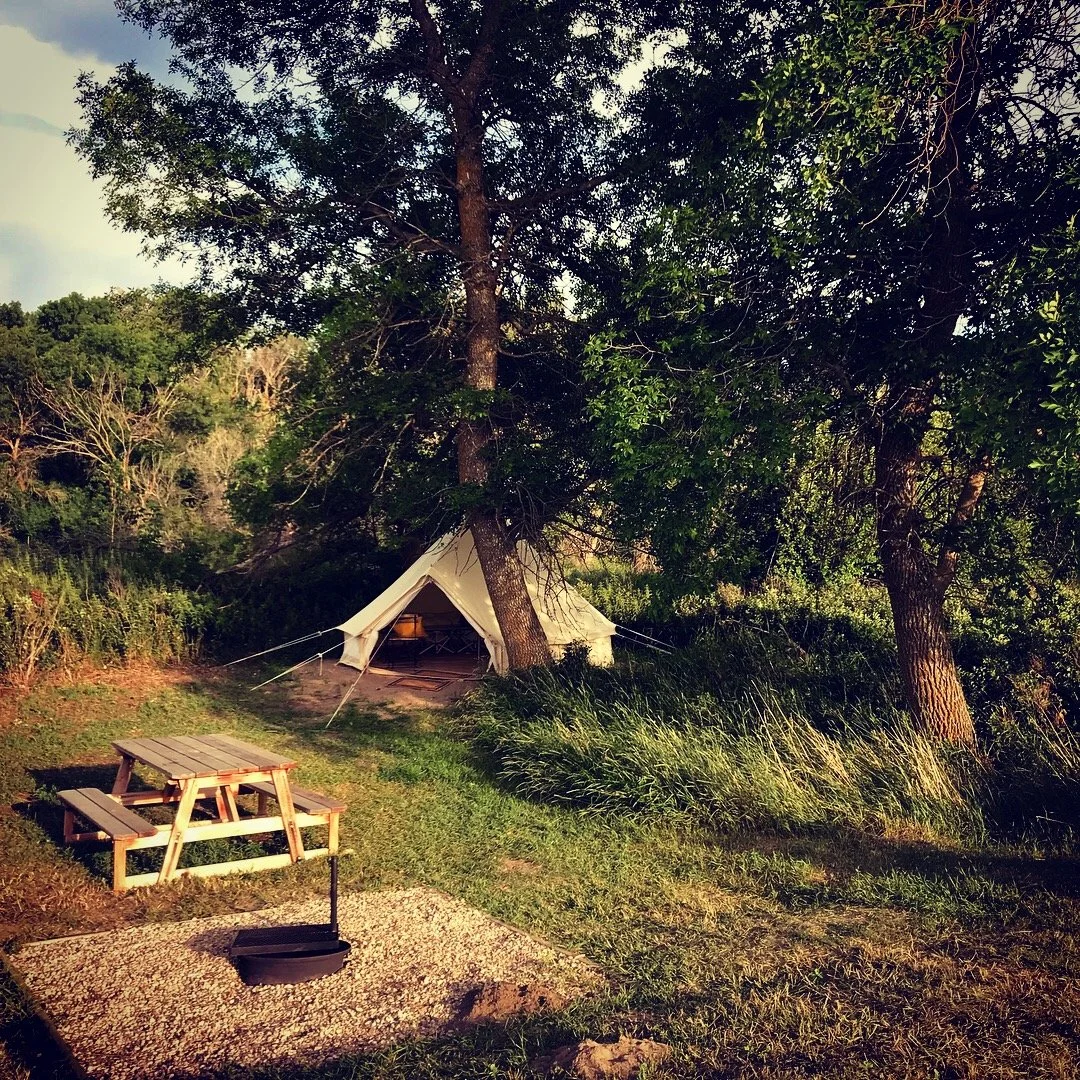 A camping scene with a canvas tent set up between two trees. There is a picnic table and a fire pit with a skillet placed nearby, surrounded by grass and trees.