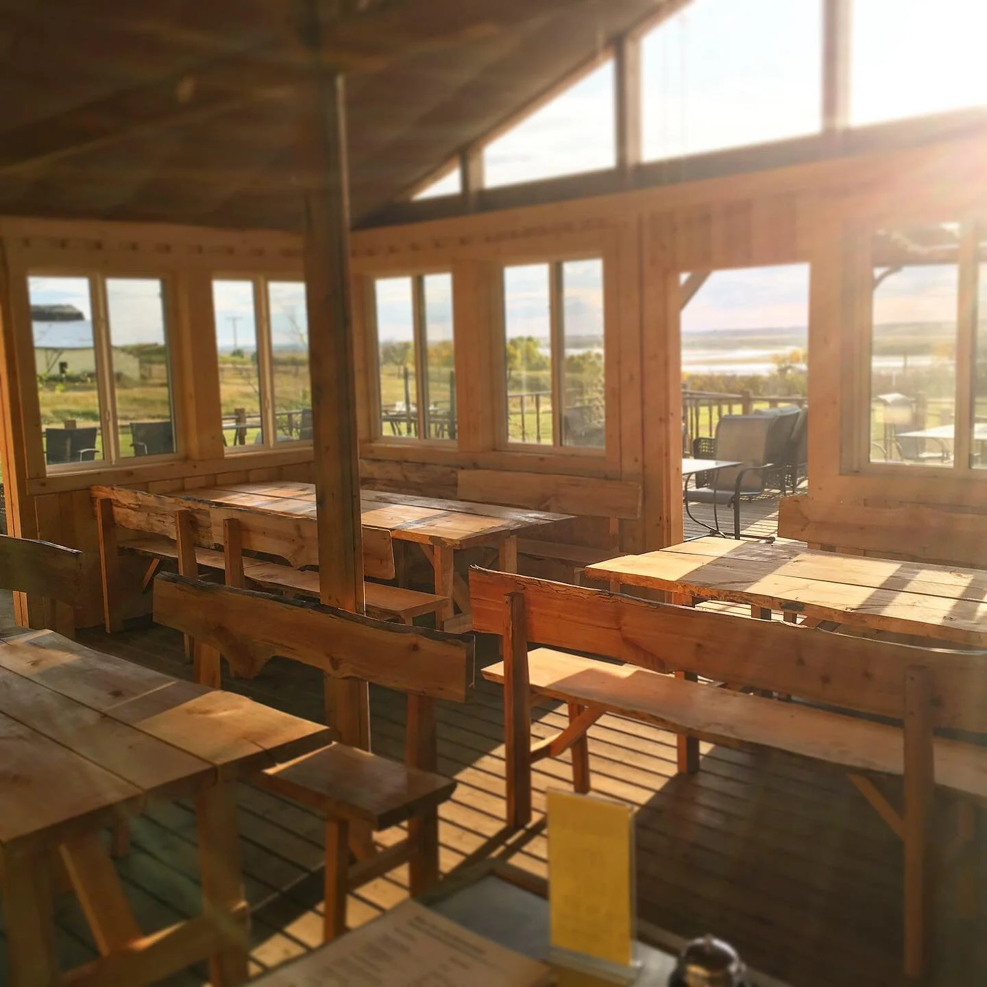 Interior of a wooden restaurant or cafe with tables and chairs, large windows showing an outdoor landscape with fields and a body of water, sunlight streaming in.