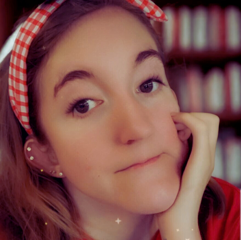 A young woman with long, light brown hair, wearing a red checkered headband, resting her face on her hand. She has multiple earrings and is in front of a bookshelf with books.