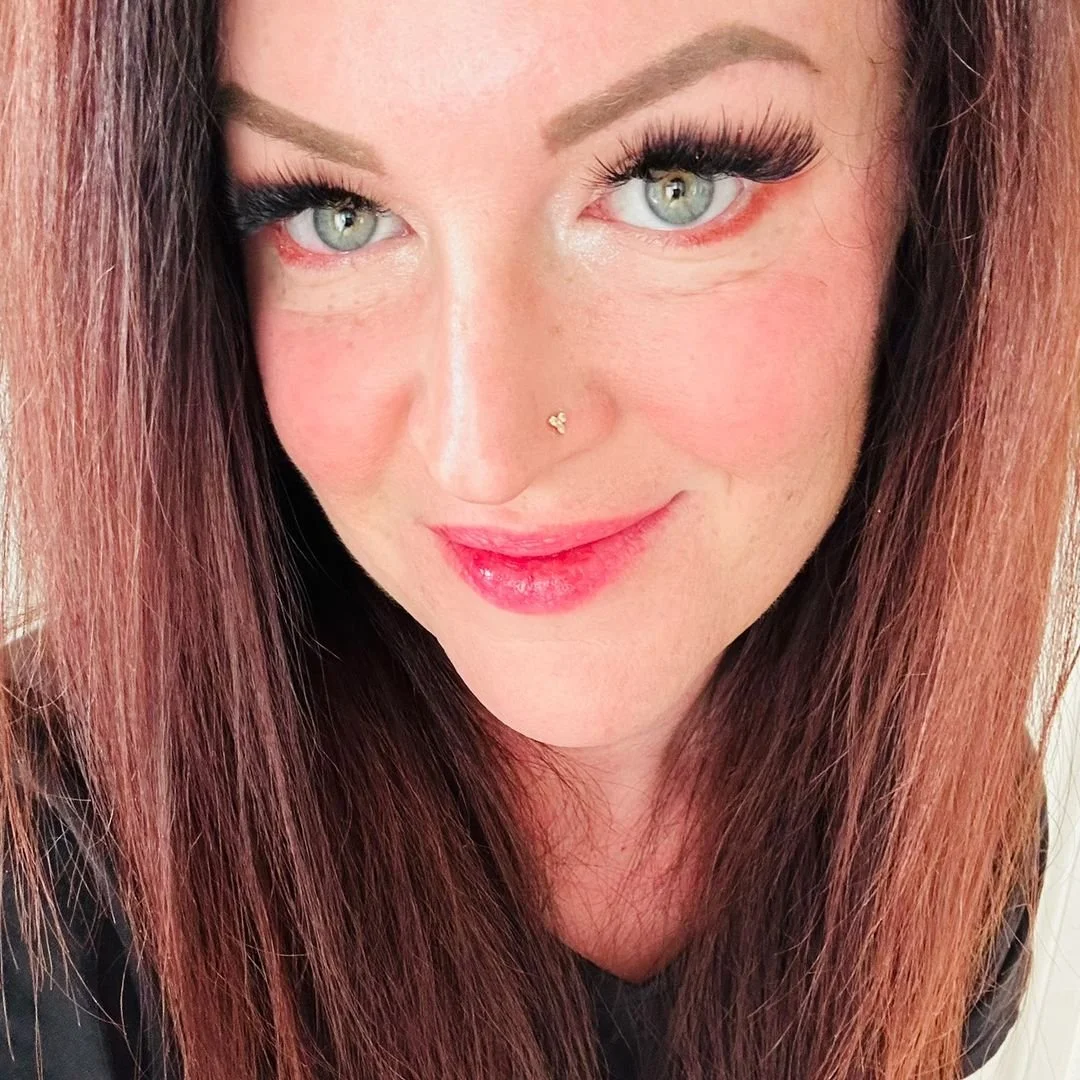 Close-up of a woman with green eyes, long reddish-brown hair, makeup, and a nose piercing.