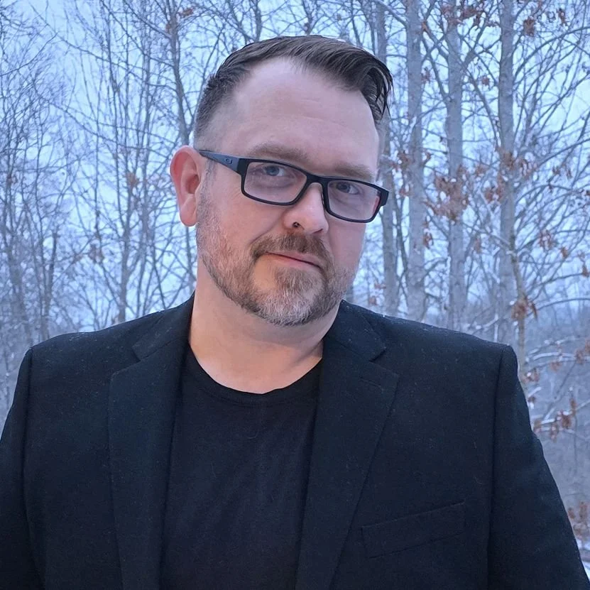 A man with short brown hair, glasses, and a beard, wearing a black blazer over a black shirt, standing outdoors in front of leafless trees with a wintery sky in the background.
